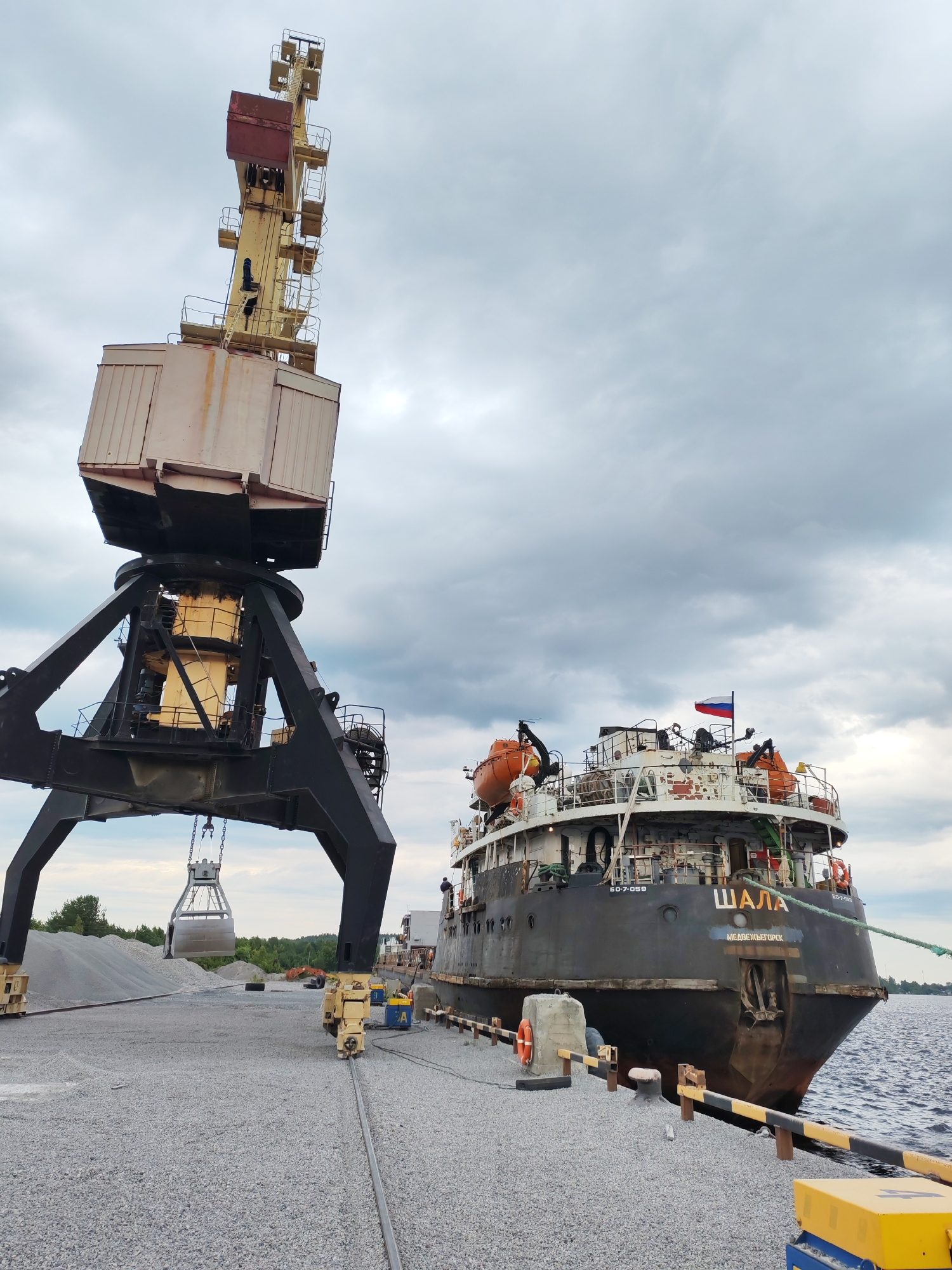 In Shalsky, Russia, a large industrial harbor scene unfolds under a cloudy sky. A massive yellow and grey gantry crane dominates the left side of the frame, its heavy lifting equipment poised over a stockpile of gravel. Below the crane, a ship, identified by the name "ШАЛА" (Shala) and "МЕДВЕЖЬЕГОРСК" (Medvezhyegorsk) painted on its stern, is docked at a pier. The ship flies the Russian flag and has orange life preservers visible on its deck. The pier is covered in gravel, with yellow and black striped bollards and a concrete structure with a life preserver on it. The water of a river or bay is visible to the right of the ship. The overall atmosphere is one of industrial activity and maritime operations.