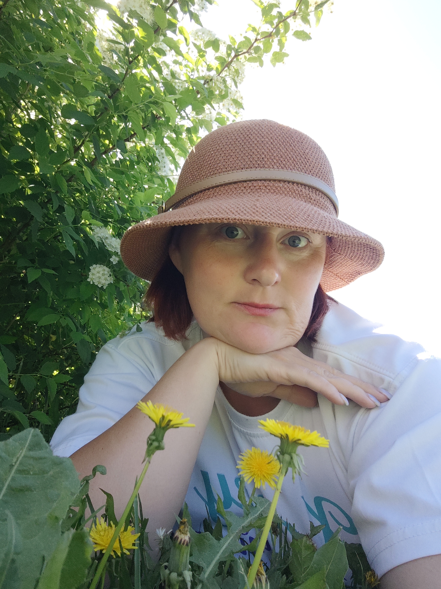 A woman wearing a brown hat and a white t-shirt with a blue design is sitting amongst dandelions in an area with green foliage and white flowers, possibly in the city of Фоки, Russia. She is resting her chin on her hand and looking at the camera. The background features sunlight and vegetation.