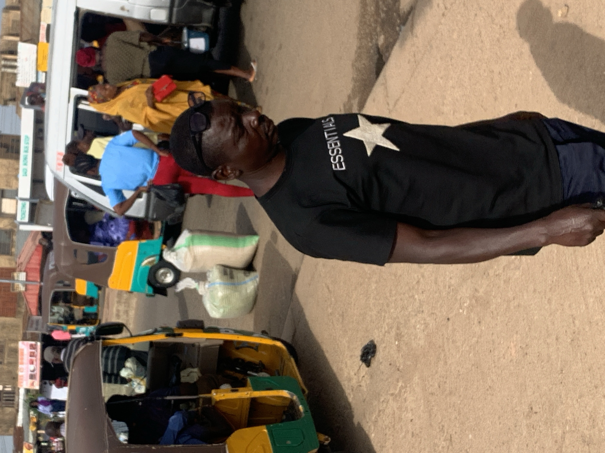 In Gura Top, Nigeria, a man stands in the foreground wearing a black t-shirt with "ESSENTIALS" and a white star printed on it, paired with dark blue pants. He has sunglasses perched on his head and his gaze is directed downwards. Behind him, a yellow and green auto-rickshaw is parked on the right side of the frame. To the left of the auto-rickshaw, there are two large sacks of goods, one light green and the other off-white. Further in the background, other people are visible near a white minibus, some boarding and others standing. The environment appears to be a busy street or market area with buildings visible in the distance.