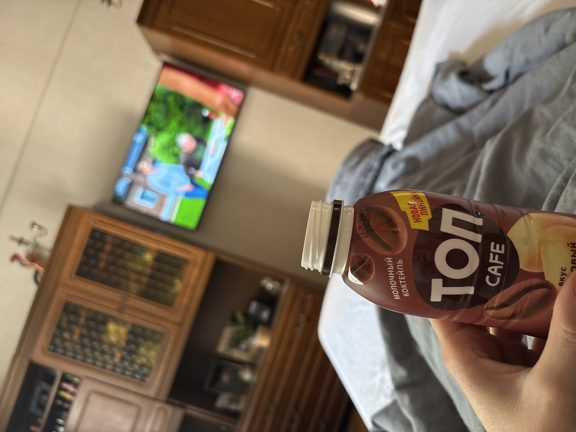 The image captures an indoor scene, primarily focusing on a person's hand holding a brown plastic bottle of a beverage. The bottle is labeled "ТОП CAFE" (TOP CAFE) and "МОЛОЧНЫЙ КОКТЕЙЛЬ" (MILK COCKTAIL), adorned with coffee bean illustrations. Visible text on the bottle also includes "НОВАЯ ЛИНИЯ!" (NEW LINE!) and partially visible "ВКУС КОФЕЙНЫЙ" (COFFEE FLAVOR), along with dates "13.05.2023" and "12.09.2023" near the cap.
In the background, a television screen displays an outdoor scene, where a person with light hair, wearing a light blue top, is visible, possibly sitting on a green surface next to blue elements. Surrounding the television are wooden cabinets or shelving units with glass doors, suggesting a classic or traditional style of furniture. Another wooden cabinet unit is present further to the left.
The foreground features a white and grey bed or bedding, indicating a relaxed, residential setting. The hand holding the bottle has fair skin. The overall scene suggests someone is enjoying a drink while potentially watching television, likely during the daytime given the ambient light. The location context provided is Minsk, Belarus.