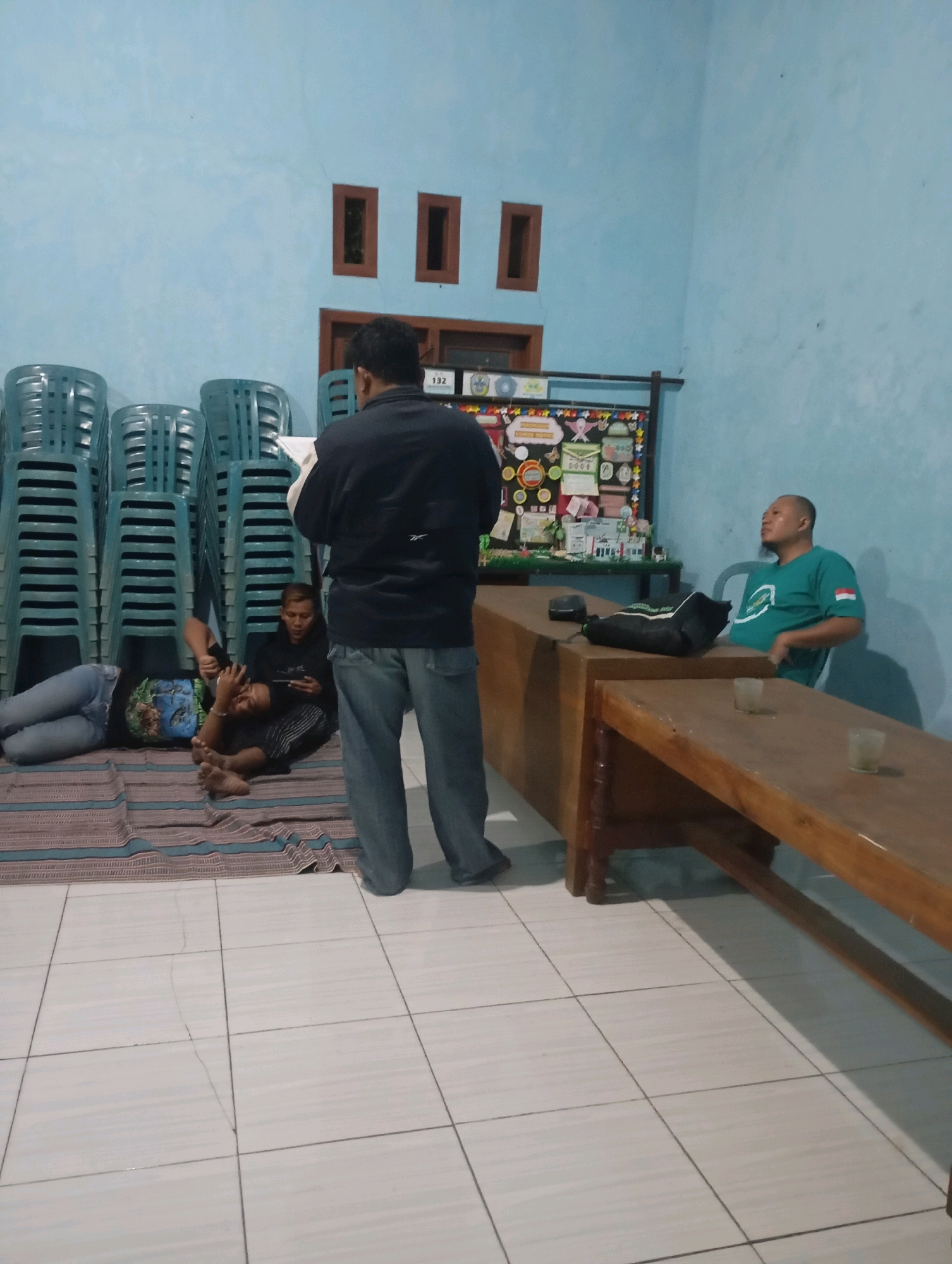 The image depicts an indoor scene, likely a community hall or meeting room, in Ketepung, Indonesia. The walls are painted a light blue, and the floor is tiled with large white square tiles, some of which show cracks. In the background, stacks of plastic chairs are neatly arranged.
Four individuals are visible. In the center foreground, a man in a dark long-sleeved shirt and blue jeans stands, facing away from the camera and appearing to read from a document or book. To his left, two people are seated on a mat on the floor. One, wearing a black hoodie, is looking down at a tablet. The other, lying down, is wearing jeans and a dark t-shirt. To the right of the standing man, a man in a green t-shirt with a flag emblem on the sleeve is seated in a chair. He is looking upwards and to the right.
A large wooden table occupies the right side of the frame, with a smaller wooden table or bench in front of it. A dark bag with a white stripe rests on the larger table. The room is sparsely decorated, with a bulletin board displaying various papers and items on the back wall. The overall atmosphere suggests a casual gathering or meeting.