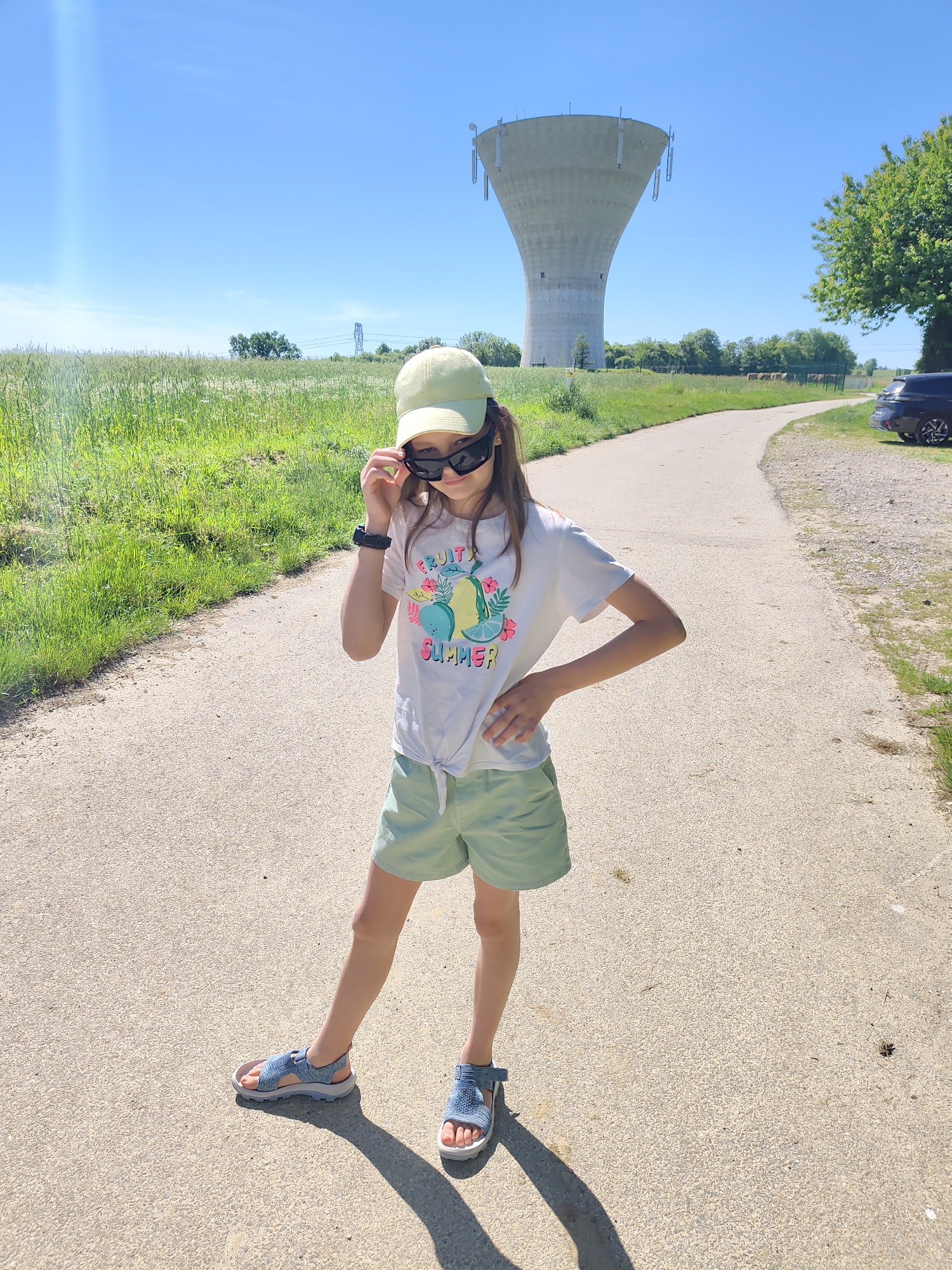 A young girl stands on a paved path, wearing a light green cap, sunglasses, a white T-shirt with the words "FRUIT SUMMER," light green shorts, and blue sandals. She holds the sunglasses on her face with her left hand and places her right hand on her hip. In the background, a water tower is visible, marking the location as Chaponnay, France. A grassy field is to the left of the path, and a road with a dark vehicle parked on the side is on the right. The sky is clear and blue.