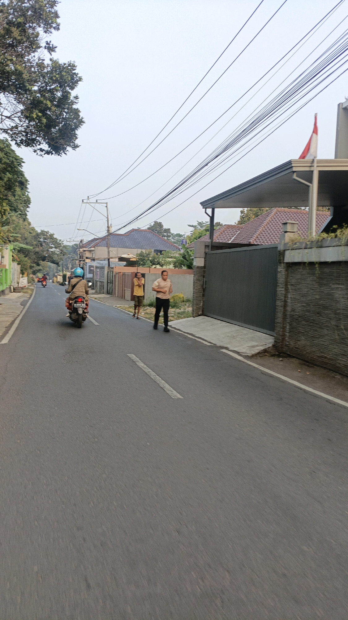 The image shows a street scene in Salatiga, Indonesia. A person wearing a light brown jacket and blue helmet is riding a motorcycle on the left side of the road. The motorcycle has a red license plate. Further down the road, two men are standing on the sidewalk on the right side of the street. One man is wearing a yellow shirt and shorts, and the other is wearing a light brown shirt and dark pants. A large gray metal gate is visible behind them. Houses with red tile roofs are visible in the background. Overhead, a complex network of power lines stretches across the sky. An Indonesian flag, red and white, is partially visible on a pole above the gate. The road is paved with asphalt and has a white dashed line marking the lane. Trees and greenery are present on both sides of the street. The sky is overcast.