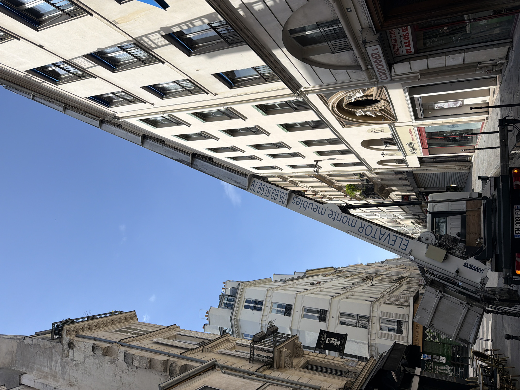 The image portrays a vibrant urban street scene, looking upwards towards the facades of several multi-story buildings under a bright, clear blue sky. The setting strongly suggests a Parisian street, characterized by light-colored, classic Haussmannian architecture featuring numerous windows, some with decorative wrought-iron railings and shutters.
The central activity involves a white moving truck, equipped with a long, extended "elevator" arm (a furniture lift), reaching high up towards the upper floors of a building on the left side of the street. This indicates a furniture moving operation is in progress, either for delivery or removal of items. The truck's license plate, "FM-309-FO," is visible, as is the brand "Böcker" on the lift mechanism.
Bright daylight illuminates the scene, casting sharp shadows from the buildings and suggesting a sunny day, likely morning or afternoon. The street level reveals various commercial establishments. Visible text on the lift's arm reads "ELEVATOR monte meubles" alongside a phone number "06.99.81.99.79". Shop signs include "CORDONNER L" (likely a cobbler), "SERRURERIE PLOM Dépannage rapid" with a phone number "01.42.46.03" (a locksmith and plumber offering rapid repairs), and "Centre de Culture Chinoise." Further down the street, a banner for "Au Vert BAR - RESTAURANT" is displayed. Stacked outdoor tables and chairs in the foreground hint at a café or restaurant. The scene captures a functional moment in a bustling city environment, combining historical architecture with modern urban services.
