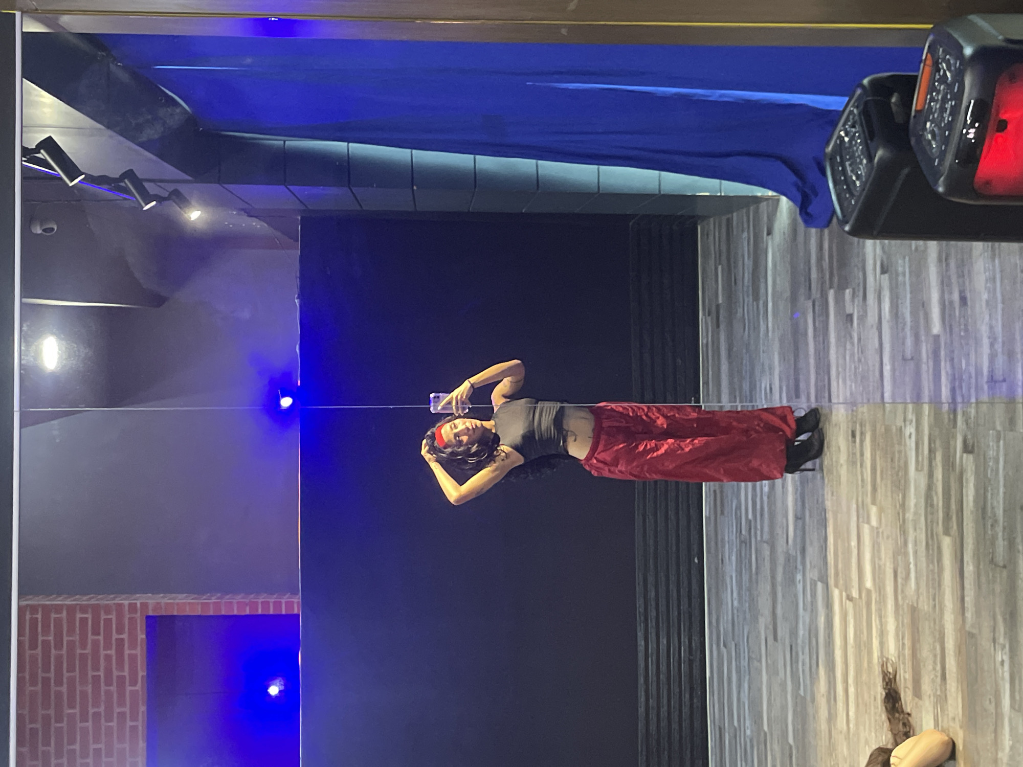 A woman is taking a mirror selfie in what appears to be a dance studio. She is wearing a grey tank top, red satin pants, and black heeled boots. She has a red headband in her curly dark hair and is posing with her hands on her head. The studio has a dark blue wall in the background with blue lights. To the right, there is a large blue curtain and some speakers are visible on the floor. The floor is made of light-colored wood planks. The image does not provide any specific information or context to place it in Pampatar, Venezuela.