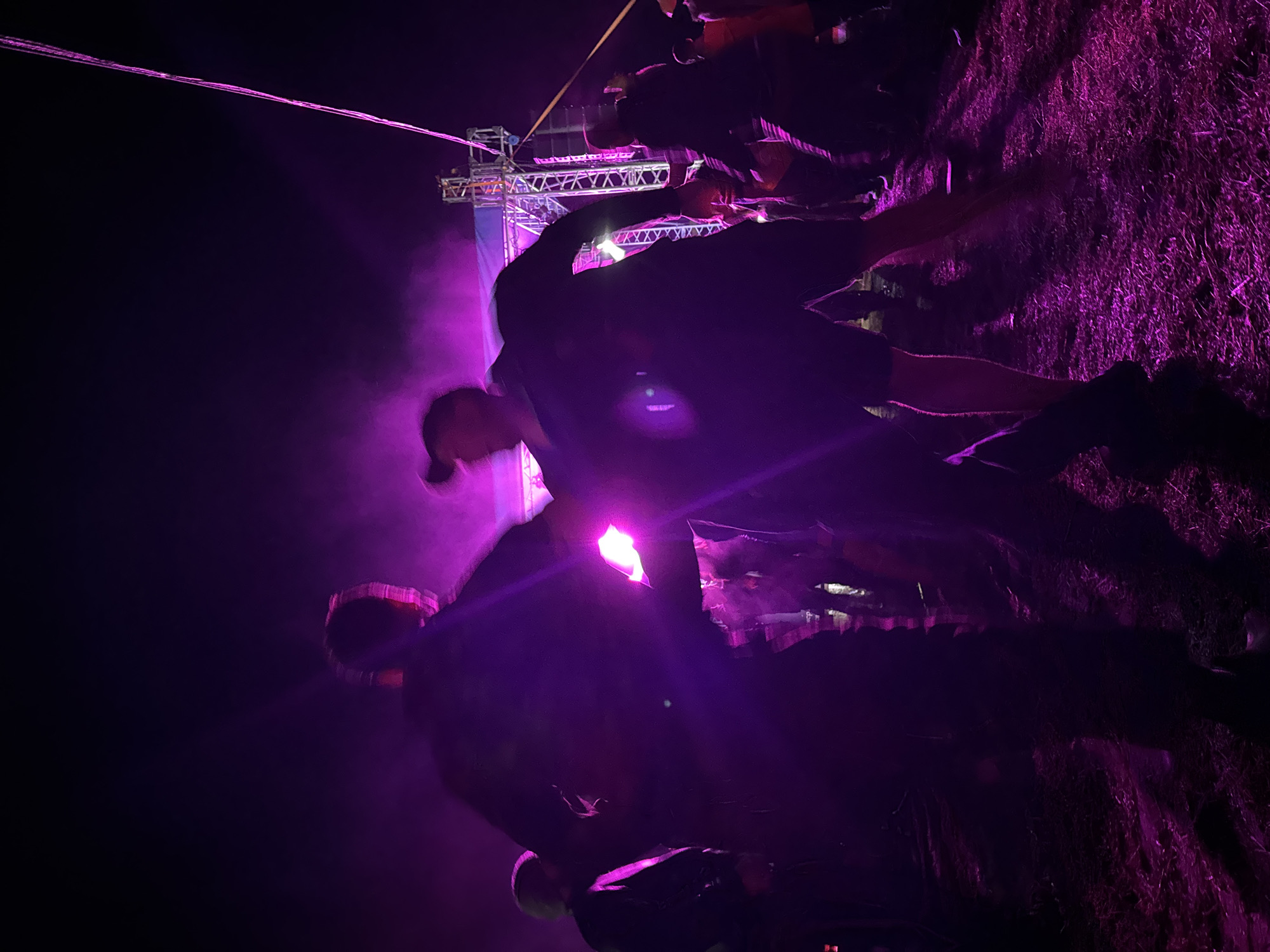 An outdoor nighttime scene is depicted at an event in Большая Кивара, Russia, characterized by strong purple artificial illumination. In the foreground, two individuals are largely silhouetted and appear to be in dynamic motion or dancing, facing each other. The person on the right is visible wearing a dark top and shorts, with their hair styled upwards, and arms in motion. Other silhouetted figures form a crowd in the mid-ground and background, indicating a public gathering. To the right, a metal truss structure, consistent with stage support or a sound system, is present. A prominent, thin pink line or cable extends diagonally from the upper right of the frame. The ground surface is unpaved, possibly dirt or sparse grass, reflecting the purple light. A atmospheric haze or smoke is visible throughout the illuminated areas.
