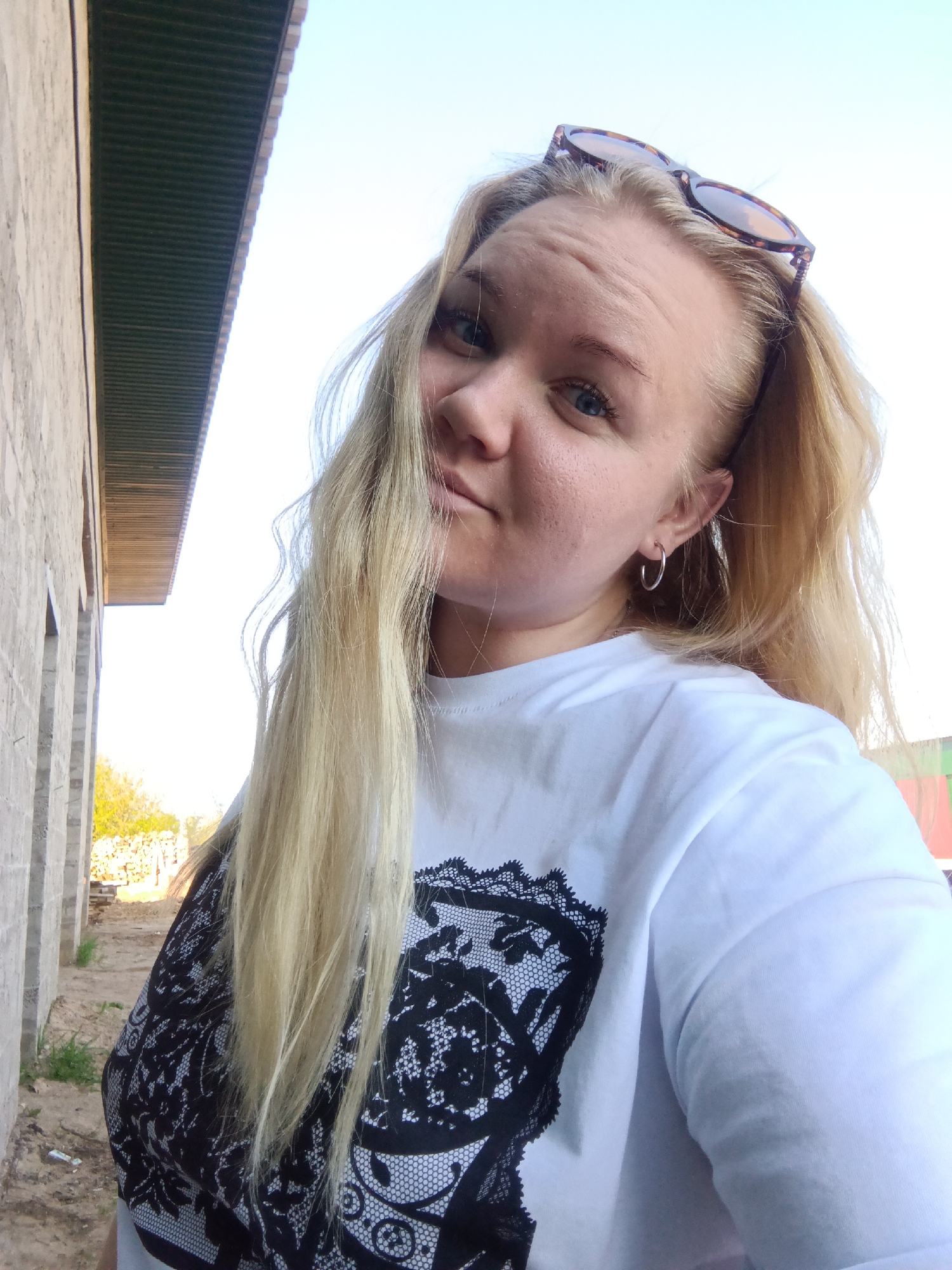 A selfie, taken from a low angle, shows a blonde woman with blue eyes. She has sunglasses resting on her head and is wearing hoop earrings. She has on a white T-shirt with a large black lace print. The background includes a light blue sky and a section of a grey brick building with a green roof overhang. The scene appears to be outdoors, possibly near the building. Based on the location, this may be taken in the city of Фоки, Russia.