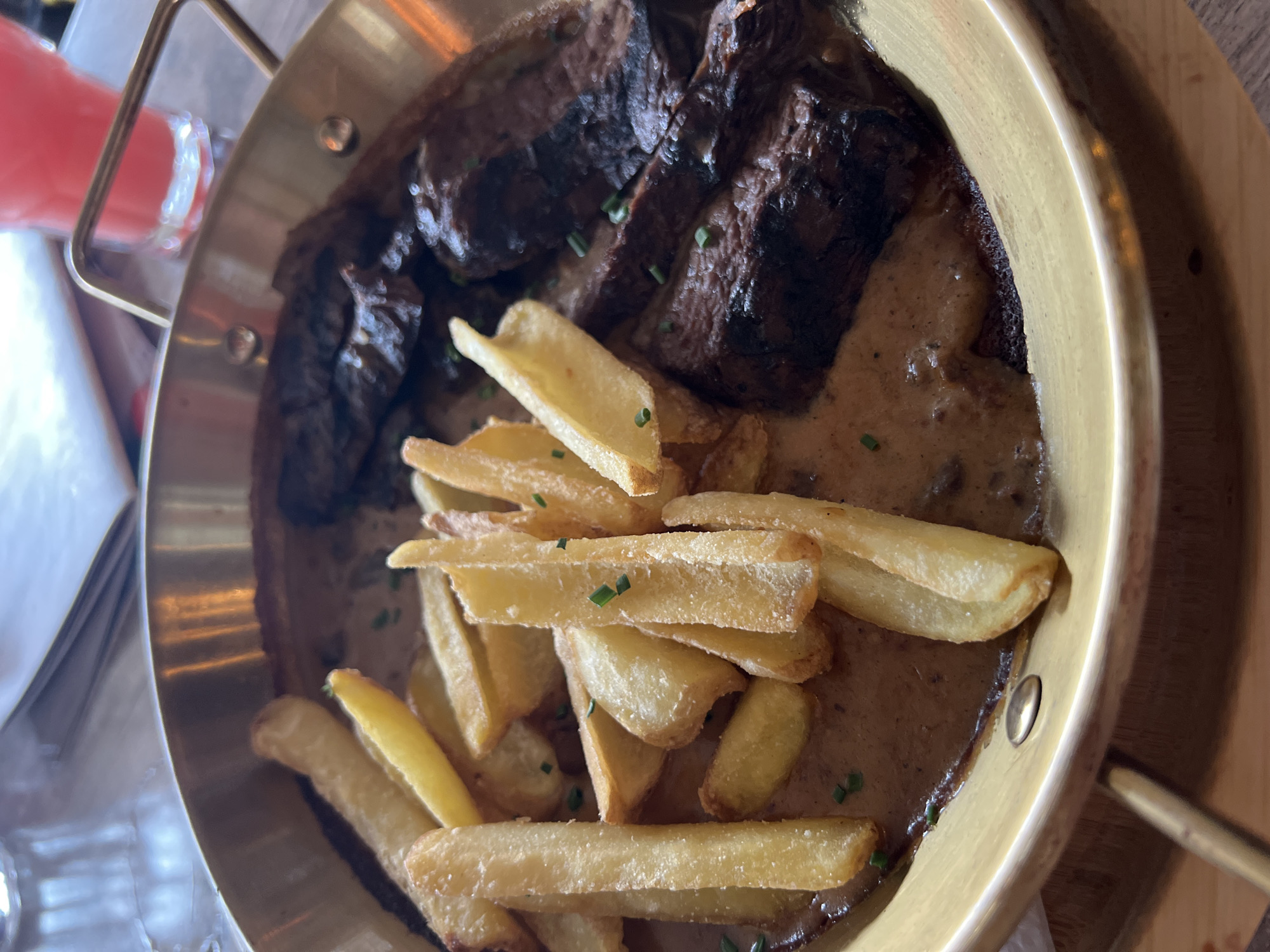 The image depicts a close-up, elevated view of a prepared meal served in a brass-colored metal pan with a handle. The pan contains several pieces of dark brown, grilled or pan-seared meat, likely steak, alongside a substantial portion of golden-brown french fries. Both the meat and fries are coated in a light brown, creamy sauce, which fills the bottom of the pan. Chopped green chives or similar herbs are sprinkled as a garnish over the food. The pan rests on a light brown wooden table surface. In the blurred background, to the upper right, a glass containing a bright red beverage is visible. The scene is set within a restaurant in Sofia, Bulgaria. No individuals, explicit actions, or direct interactions are visible.