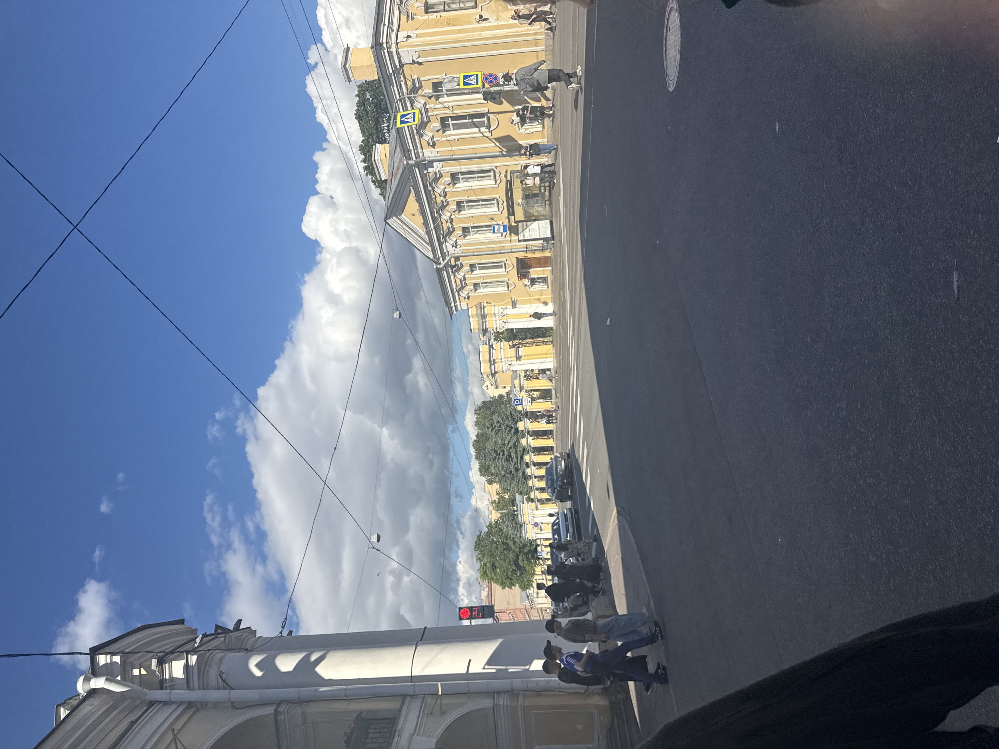 A street scene in Saint Petersburg, Russia, on a sunny day with scattered clouds. The image features several people, cars, and buildings typical of an urban environment. On the left side of the frame, a tall, light-colored building with an arched entrance dominates the foreground. Adjacent to this, a group of young people is visible, some walking and others standing near a crosswalk. A red traffic light is illuminated above them, with a digital timer showing "24."
Across the street, a long, yellow building with ornate white detailing lines the right side of the image. This building appears to be a historic structure with multiple windows and a prominent facade. Several individuals are seen walking along the sidewalk in front of this building, and a few cars are present on the road, some of which are moving. Pedestrian crossing signs with yellow backgrounds are visible, indicating a marked crosswalk. The street surface is asphalt, and overhead power lines crisscross the sky. The overall impression is that of a bustling urban intersection, likely in a historical district of the city.