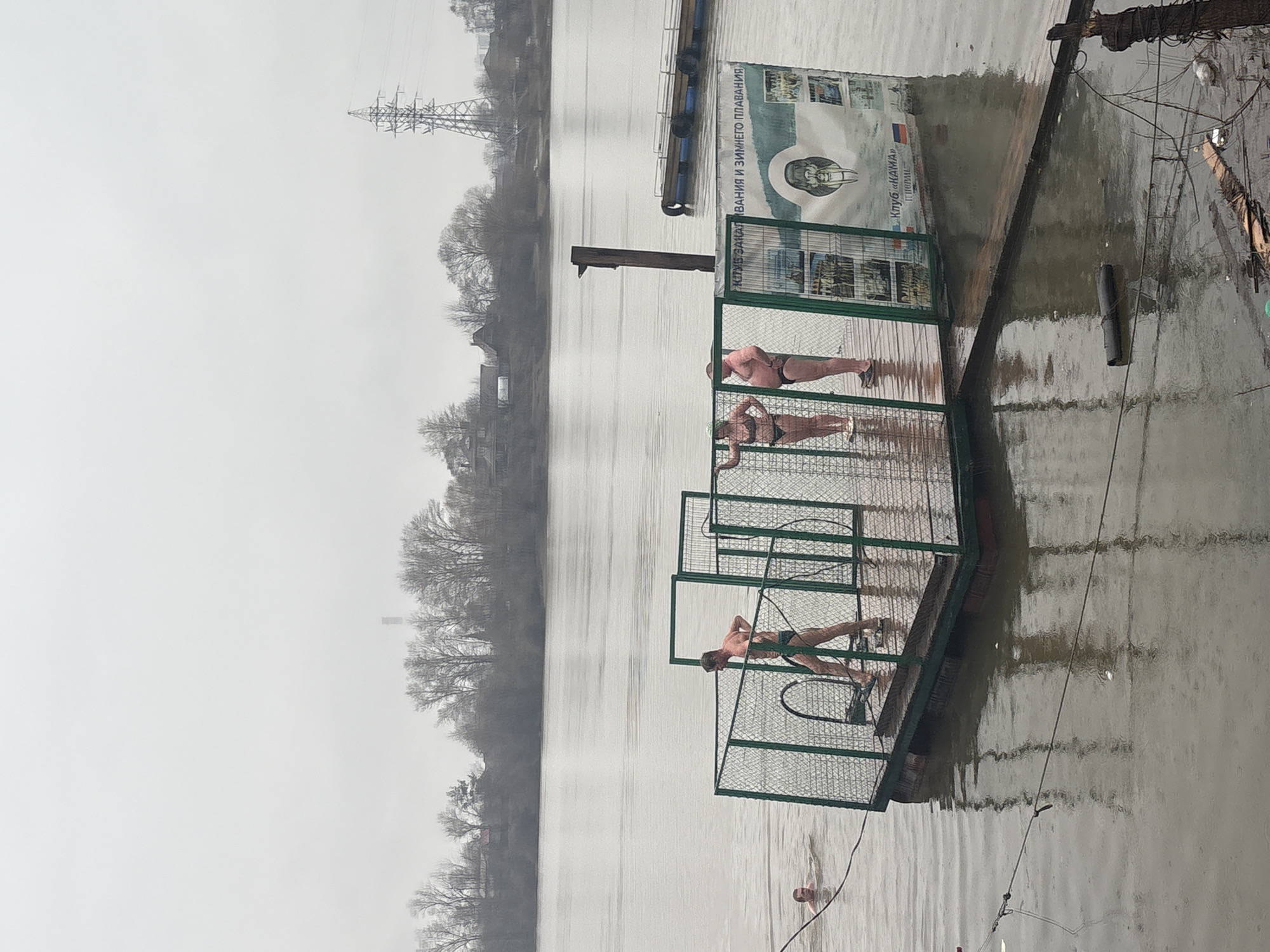 The image shows three people standing on a small fenced platform extending into a body of water, possibly the Kama River. The people appear to be dressed in swimming attire. There is a sign on the dock near the platform that is likely related to a swimming club. The environment appears overcast and the water is grey. In the background there are buildings, trees, and an electrical transmission tower. The city is Perm, Russia.