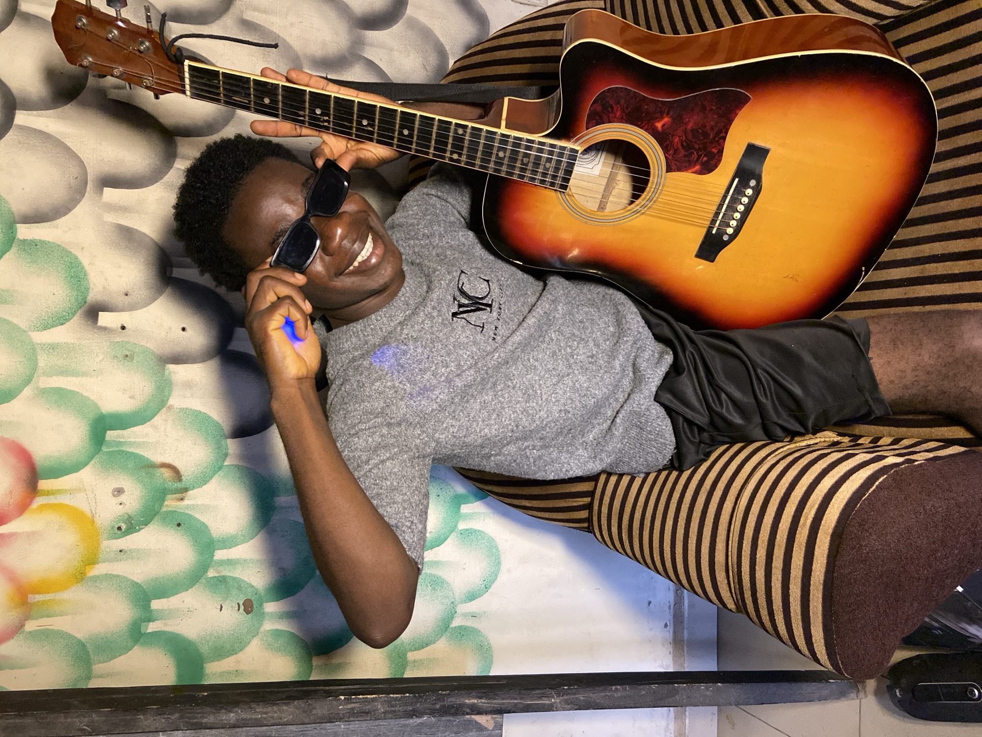 An indoor, portrait-oriented image captures a young Black man lying on his back, smiling broadly towards the camera. He is positioned diagonally, with his head in the upper left of the frame. He wears black, square-framed sunglasses, a grey long-sleeved t-shirt, and black shorts. His short, dark, curly hair is visible. In his left hand, he holds a small, glowing blue object, possibly a light stick or vape pen.
An acoustic guitar with a sunburst finish (yellow-orange fading to brown) is placed across his body, held by his right hand. The guitar's headstock, fretboard, strings, soundhole, and bridge are clearly discernible.
The man rests on what appears to be a sofa or cushion covered in a fabric with distinct horizontal brown and gold/tan stripes. Behind him, on the left side of the image, is a wall or surface decorated with an abstract pattern of overlapping, roughly circular or oval shapes in shades of light green, yellow, and hints of pink.
The man appears cheerful and relaxed, exuding a happy and playful mood. Visible text on his grey t-shirt includes stylized letters "NC" and the phrase "NEW YORK CITY." The setting is casual and informal, situated in Mararaba, Nigeria.