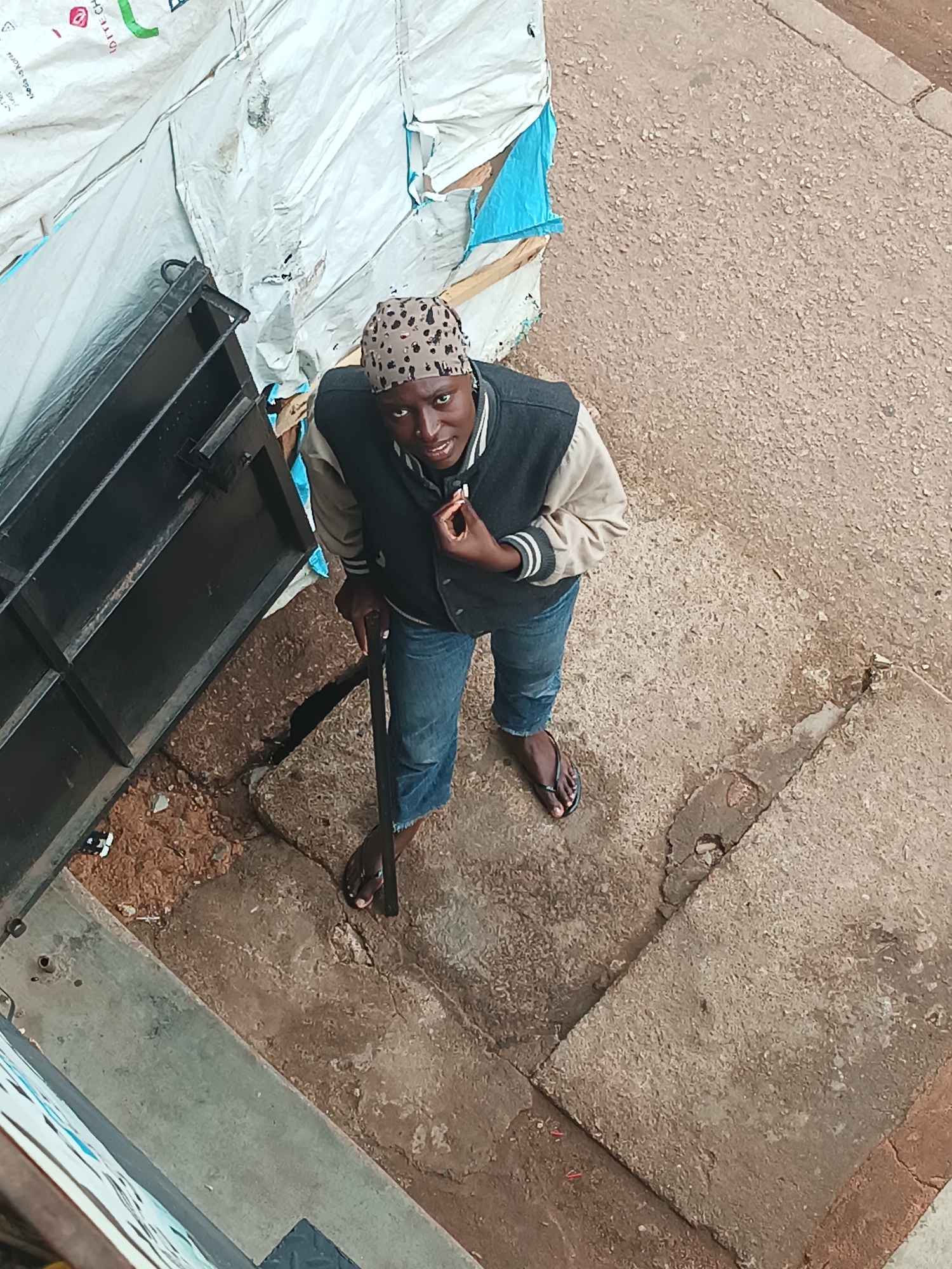 An overhead shot captures a man standing outdoors on a surface composed of rough concrete slabs and reddish-brown dirt and gravel. He is looking directly up at the camera with a subtle smile. The man is wearing a spotted or patterned head covering, a two-tone jacket with a dark body and light-colored sleeves featuring striped cuffs, and blue jeans. He is barefoot, wearing dark flip-flops. In his left hand, he holds a dark stick or pole, resting its end on the ground. His right hand is raised, holding a small, light-colored object near his mouth.
To the man's left, a large, crinkled white tarp or sheet is visible, appearing to cover or be draped over something. Faint text "Made in Korea" is discernible on the tarp, along with other partially obscured letters like "DITE CH". Next to the tarp is a dark, rectangular metal structure or frame. The scene is illuminated by natural daylight. The overall setting suggests an informal outdoor area, possibly in a market or residential street in Bukuru, Nigeria.