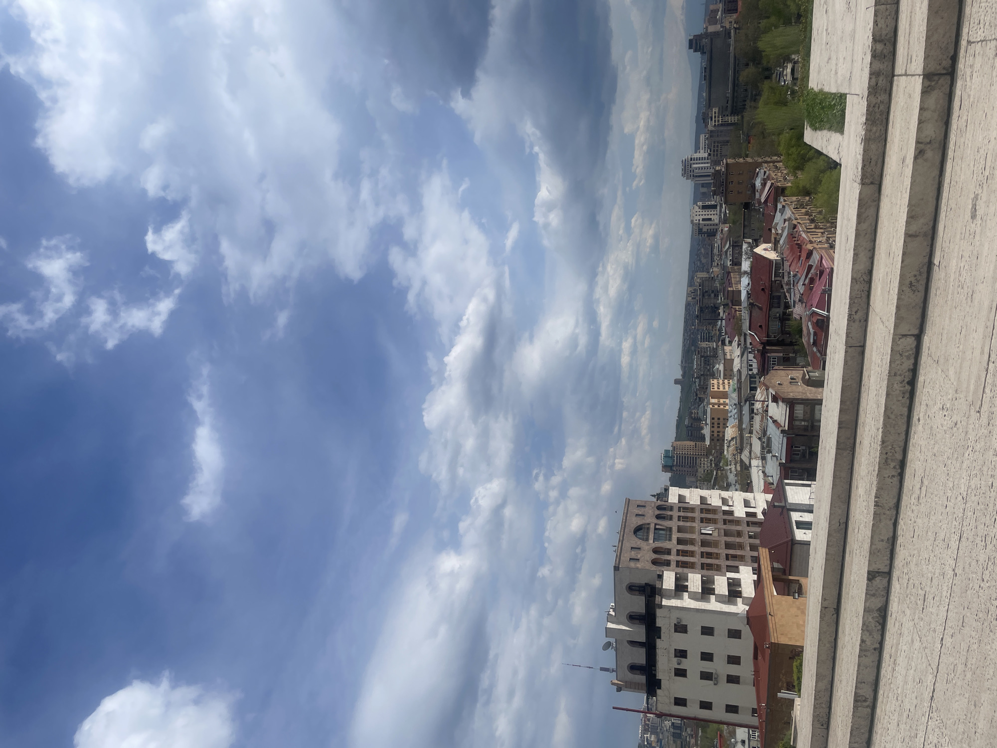 The image shows a cityscape viewed from a high vantage point, likely in Yerevan, Armenia. The foreground includes stone steps or ledges. Below, a mix of residential and commercial buildings are visible, with rooftops of varying colors and architectural styles. In the background are taller apartment buildings. The sky is filled with white and gray clouds against a blue sky. There are no people visible.