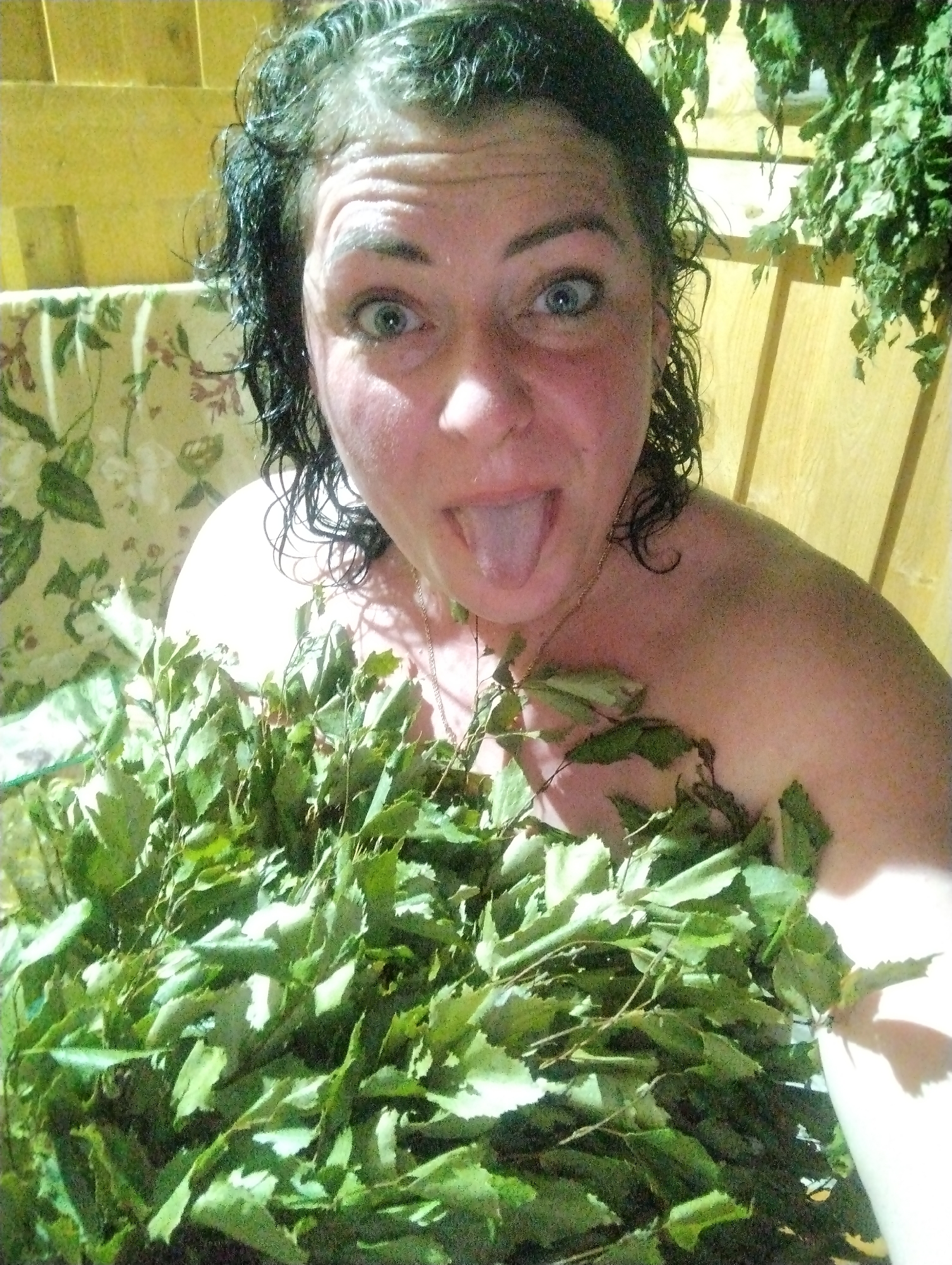 A woman with wet, dark hair and flushed skin is holding a large bunch of green leafy branches in front of her. Her eyes are wide, and she is sticking her tongue out. She appears to be in a sauna, as indicated by the wooden paneling and leafy bundles hanging in the background. A floral-patterned fabric is also visible in the background. A gold chain is visible around her neck. It's not possible to definitively confirm the location of the image as Grishanki, Russia, from the visual information alone.