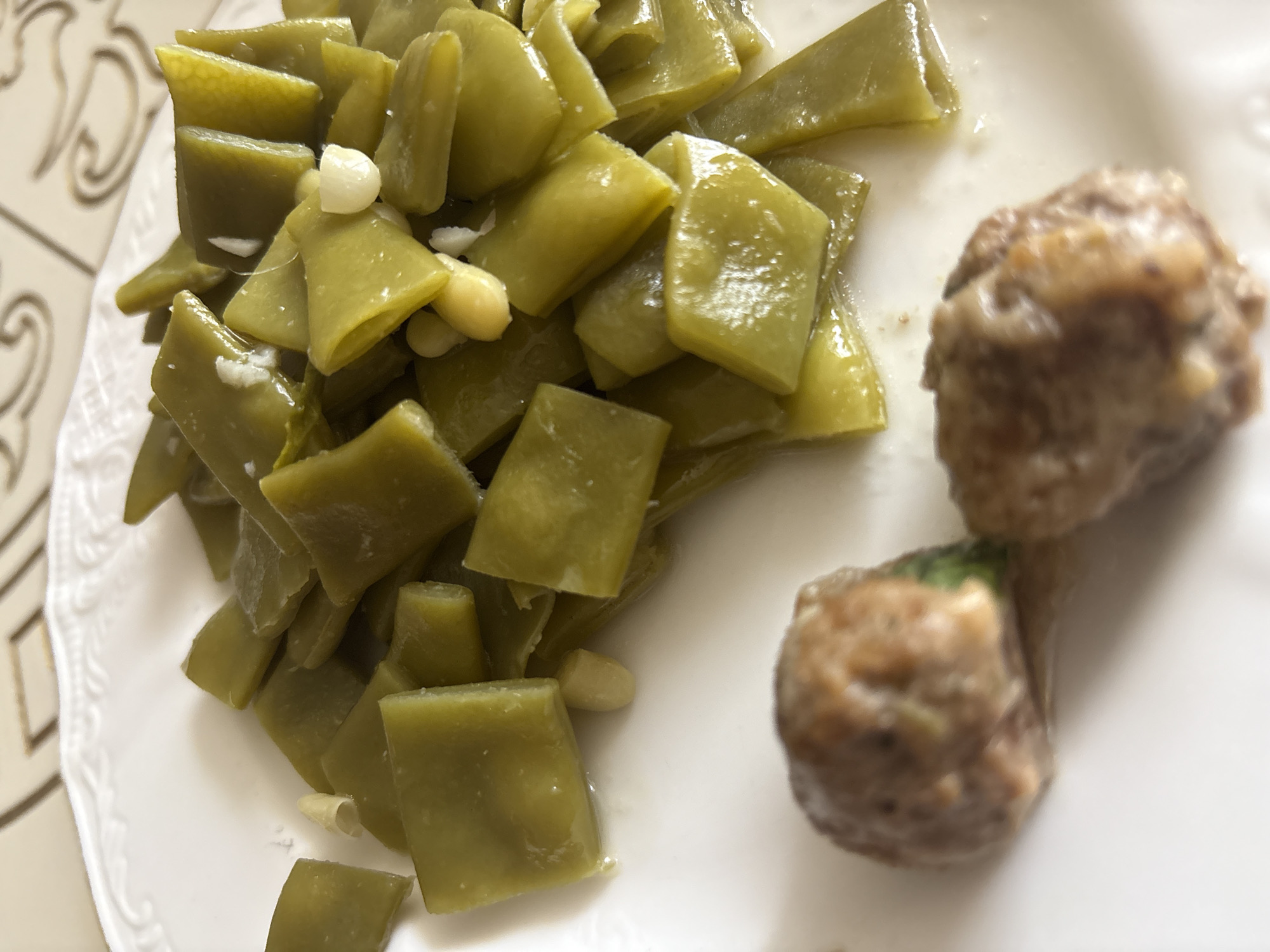 A white plate holds a serving of cooked green beans and two meatballs. The green beans are cut into diagonal pieces and appear to be seasoned with garlic. The meatballs are round and brownish-gray, with visible texture. The plate is set on a table with a light-colored, patterned tablecloth. The scene is a close-up, with the food taking up most of the frame. There is no information in the image to suggest a location in Georgiyevsk, Russia.