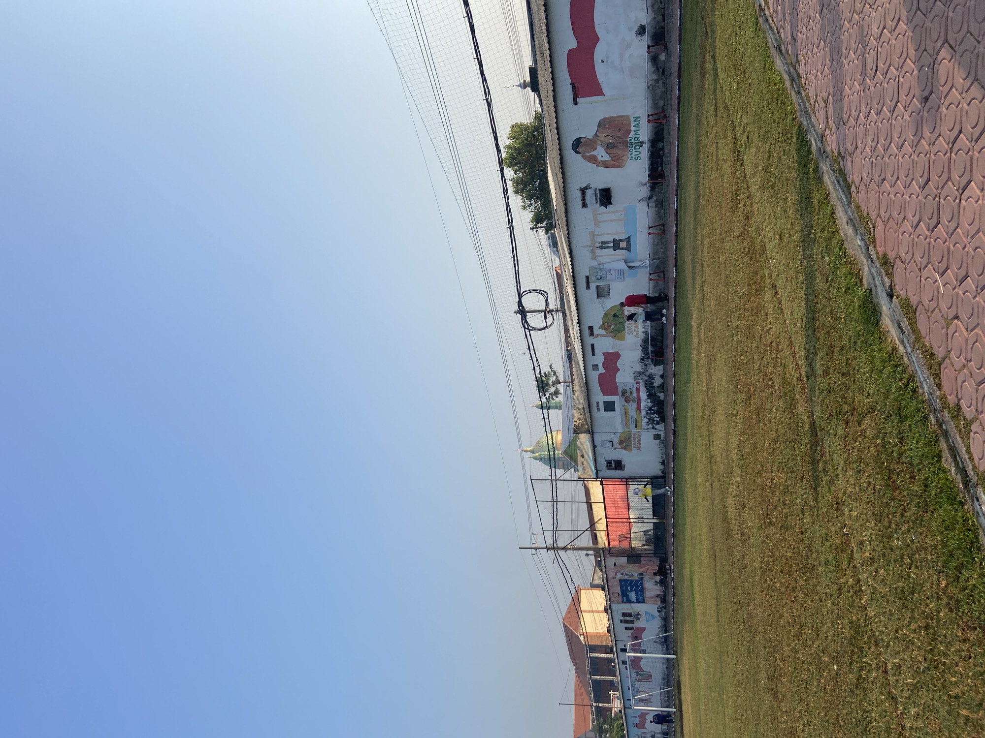 The image depicts a clear, sunny day in Sumorame, Indonesia, with a bright blue sky overhead. In the foreground, a well-maintained grassy field, likely a soccer field, stretches out. To the right, a paved walkway with a distinctive hexagonal pattern borders the field.
Dominating the background is a long, white building adorned with murals. These murals feature patriotic Indonesian themes, including large depictions of the Indonesian flag, portraits of historical figures such as "JENDERAL SUDIRMAN," and various symbolic imagery. The building itself appears to be low-rise, with a corrugated roof and several windows. A sports net, possibly for a court adjacent to the field, is strung across the top of the frame.
Several individuals are visible in the scene. Two people in red and white clothing are near the building, engaged in conversation or observation. Another person, wearing a yellow top and dark pants, is standing on the field. A few other figures are smaller and more distant, appearing to be walking or engaged in activities on or near the field.
In the distance, behind the building, glimpses of other structures can be seen, including a building with an orange roof and a building topped with a green dome, suggesting a mix of residential and possibly religious or institutional architecture. Trees are also present, adding to the outdoor setting. The overall impression is one of a community space, possibly a sports complex or a public park, that has been decorated with art and is in active use.