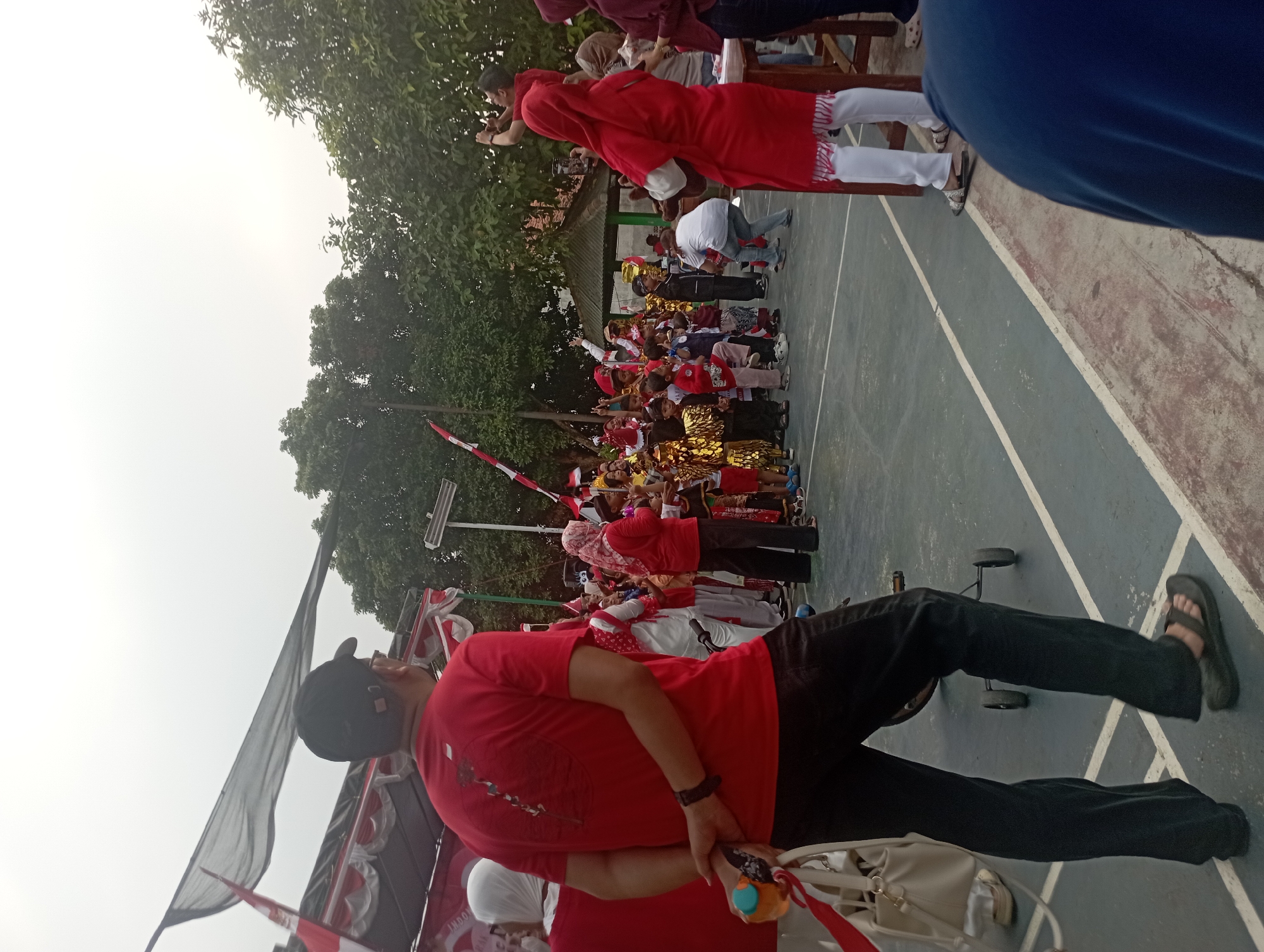 This image captures a lively outdoor event, possibly a school or community gathering, in Purwakarta, Indonesia. The scene is filled with people, predominantly children, dressed in festive red and white attire, suggesting a national celebration or holiday. Many of the children are wearing costumes, some adorned with gold accents, and some are holding small flags.
In the foreground, a man wearing a red t-shirt and black pants stands with his back to the camera, holding a blue object. He appears to be facing the group of children and adults. To his left, a white bag is visible. The ground is a light blue court with white lines, resembling a basketball or badminton court.
Behind the children, a stage or platform is set up, decorated with red and white banners. Trees and a building with a dark roof are visible in the background, indicating an outdoor setting in a populated area. The overall atmosphere seems joyful and energetic, with a sense of community participation.