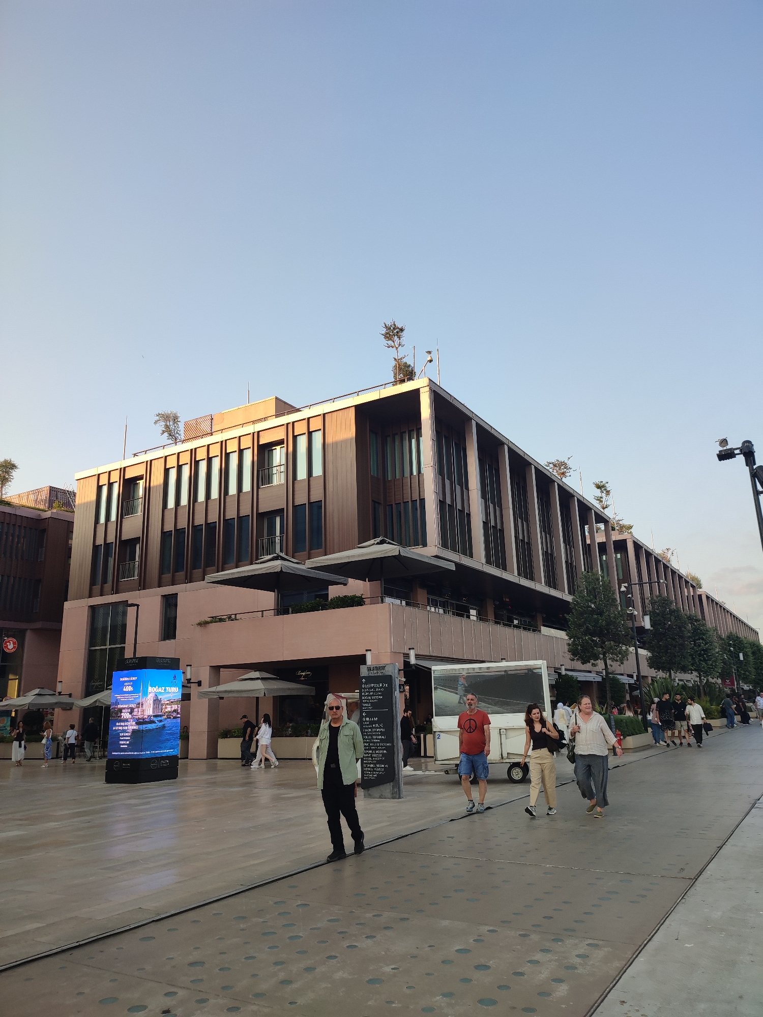 An outdoor plaza in the city of Beyoğlu, Turkey, features a multi-story building with a modern facade of wood-paneled sections and vertical window frames. The building has several levels with balconies and is topped with trees. In the foreground, people are walking across a patterned paved area. To the left, a large digital billboard displays an advertisement. Several umbrellas are set up on the ground level of the building, suggesting an outdoor seating area for businesses. Trees are planted along the side of the building. The sky is clear and pale blue, indicating daytime.