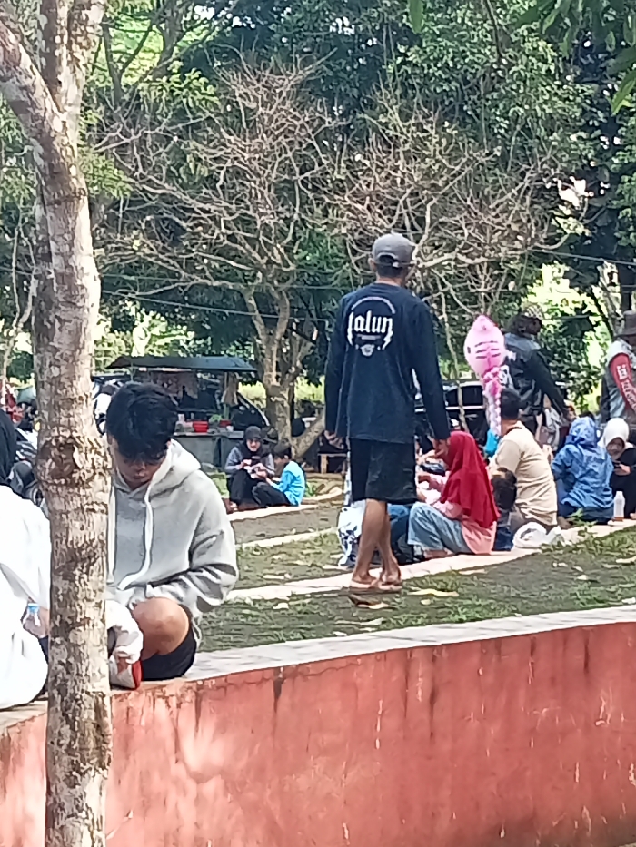 This outdoor image captures a casual gathering of people in Singaparna, Indonesia, likely in a park or public green space during the daytime. The scene is dominated by lush trees, some with dense foliage and others with bare, intricate branches, suggesting a natural environment.
In the immediate foreground on the left, a young person with dark hair, wearing a light grey hoodie and dark shorts, is seated on a low, reddish-brown concrete wall. They are looking downwards, appearing focused on something in their lap.
Centrally positioned in the mid-ground, seen from behind, is a man walking to the right. He wears a dark blue long-sleeved shirt with a white circular logo on the back that clearly reads "alun". He also wears dark shorts, a grey baseball cap, and flip-flops.
Around him and further into the background, numerous other individuals are scattered across grassy areas and concrete steps. Many are sitting on the ground, some in small groups. Notable figures include a person in a red hijab sitting on the grass, and another person holding a pink, seemingly inflatable object (possibly a balloon). In the far background on the left, under some trees, there appears to be a small stall or tent, with more people gathered nearby.
The overall atmosphere seems relaxed and informal, suggesting a leisure activity or a community gathering. The lighting indicates it is daytime, with bright conditions.