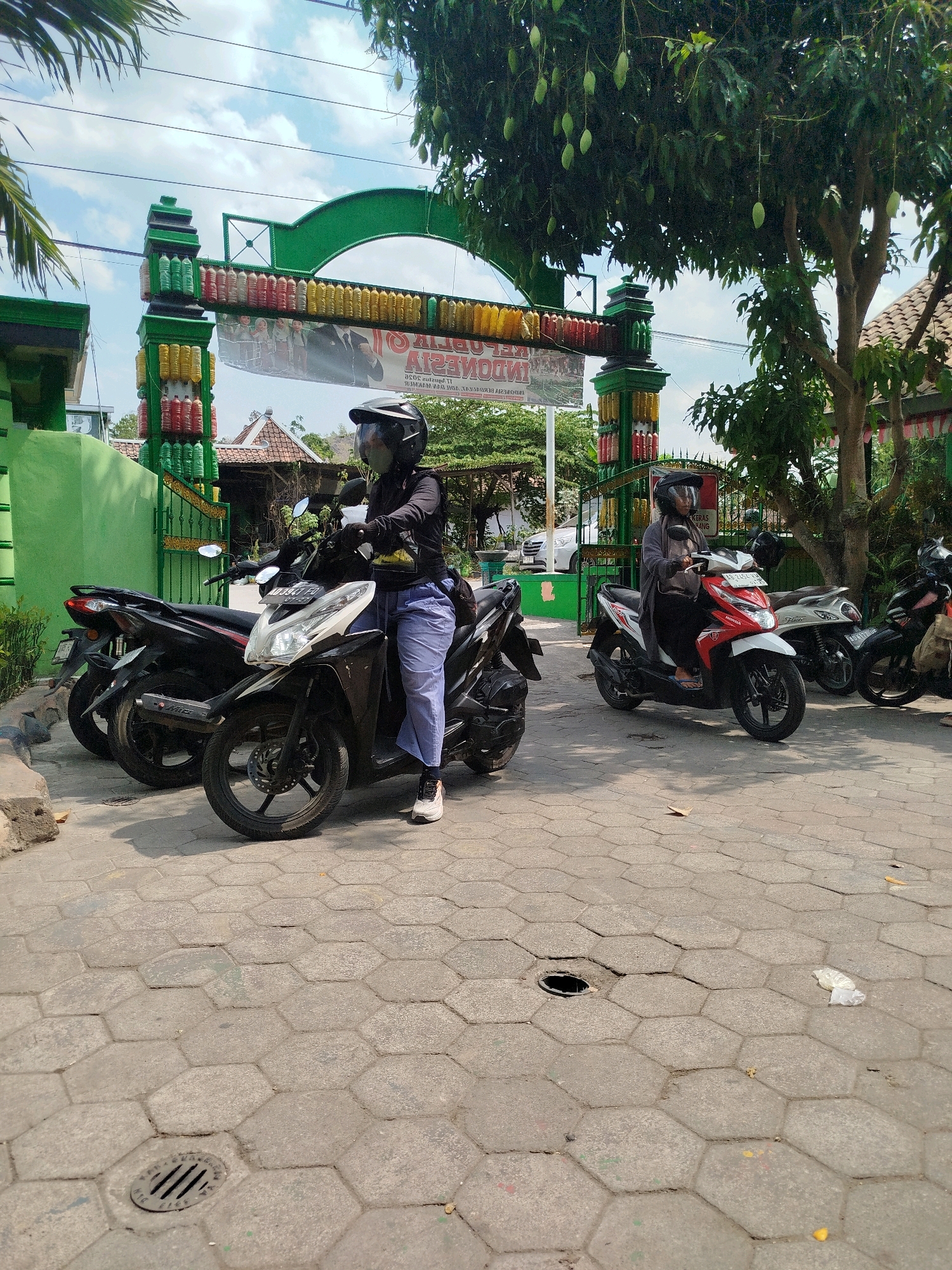 In Grogol, Indonesia, an outdoor scene depicts several parked motorcycles in front of a green archway decorated with colorful plastic bottles and a banner. The archway features a large green structure with decorative elements made from what appear to be recycled plastic bottles in red, yellow, and green. A banner with text in Indonesian is suspended across the arch. Two individuals are seated on motorcycles, both wearing helmets. One person, positioned in the foreground on a dark-colored scooter, is looking towards the viewer and wearing a black long-sleeved shirt, light blue pants, and white sneakers. Another individual is seated on a red and white scooter in the mid-ground, also wearing a helmet and dark clothing. Several other motorcycles are parked in the vicinity. The ground is paved with hexagonal concrete tiles, and a metal drain cover is visible. Trees with hanging fruit, possibly mangoes, are present in the background, along with some buildings with tiled roofs. The sky is partly cloudy.