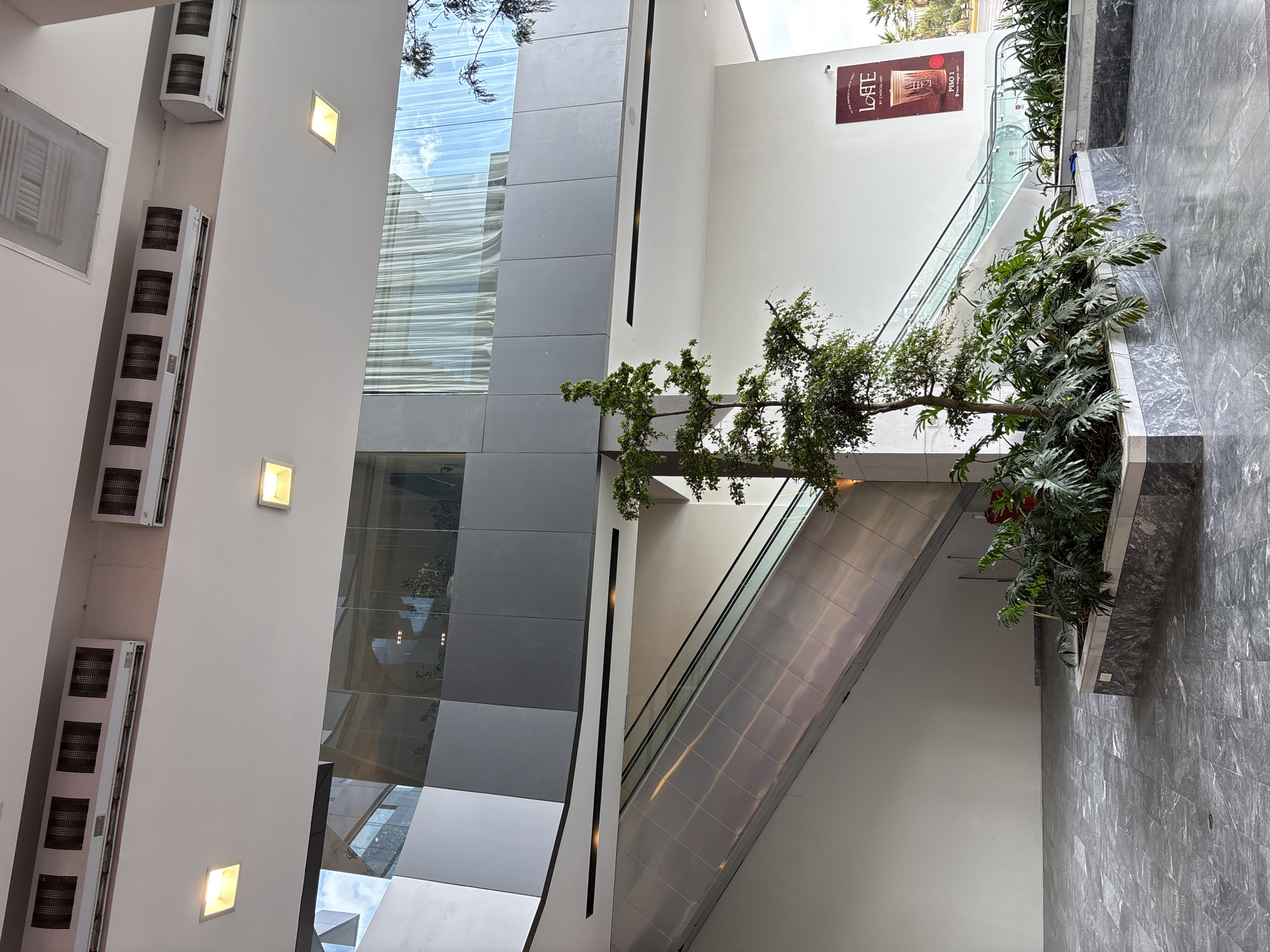 The image depicts the interior of a multi-story building, likely a shopping mall, in San Bernardino Tlaxcalancingo, Mexico. The scene is dominated by architectural features, including an escalator, a multi-story glass window, and an elevator shaft. The escalators lead towards a landing adorned with potted plants. A tree or shrub is positioned in front of the elevator shaft. On the left side of the image, a white wall is fitted with multiple air conditioning units and square light fixtures. An advertisement is visible on the upper level, featuring the words "LOVE by CHOCOLAT." The floor is tiled.