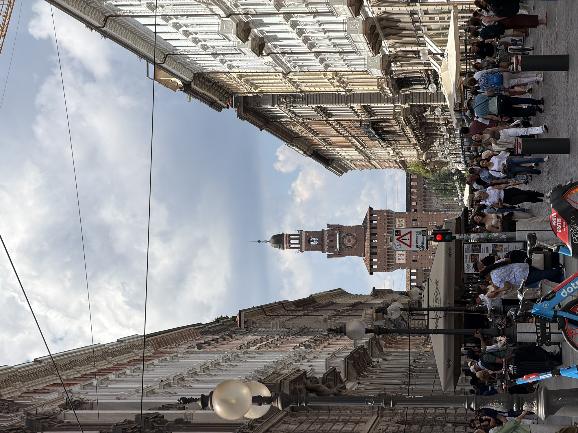 This vertical shot captures a lively street scene in Milan, Italy, with the iconic Sforza Castle's clock tower visible in the distance. The perspective is looking down a street flanked by tall, ornate buildings featuring classical architectural details, including cornices, pilasters, and decorative moldings. The sky above is bright with scattered clouds.
Numerous people populate the street, primarily gathered around outdoor seating areas belonging to cafes or restaurants. Many are seated at tables under large, light-colored umbrellas, appearing to be dining, drinking, and conversing. Others are standing or walking along the cobblestone pavement. The atmosphere suggests a casual, bustling daytime environment.
Notable objects include several streetlights with globe-shaped lamps, power lines crisscrossing the sky, and various street furniture elements. In the foreground and midground, several shared electric scooters or bicycles are parked, some in distinctive blue with "dot" or "dotty" branding, and one red and black scooter displaying "PROVA GRATIS!" (Try for free!) and a "BOOM" logo. A red traffic light is visible near the castle tower, next to a triangular warning sign with a red border and an exclamation mark, below which "0-24" is legible. Part of a "Hard Rock Cafe" sign can also be seen. The people are dressed in everyday clothing, typical for daytime urban activities. The overall impression is one of a vibrant European city enjoying good weather.