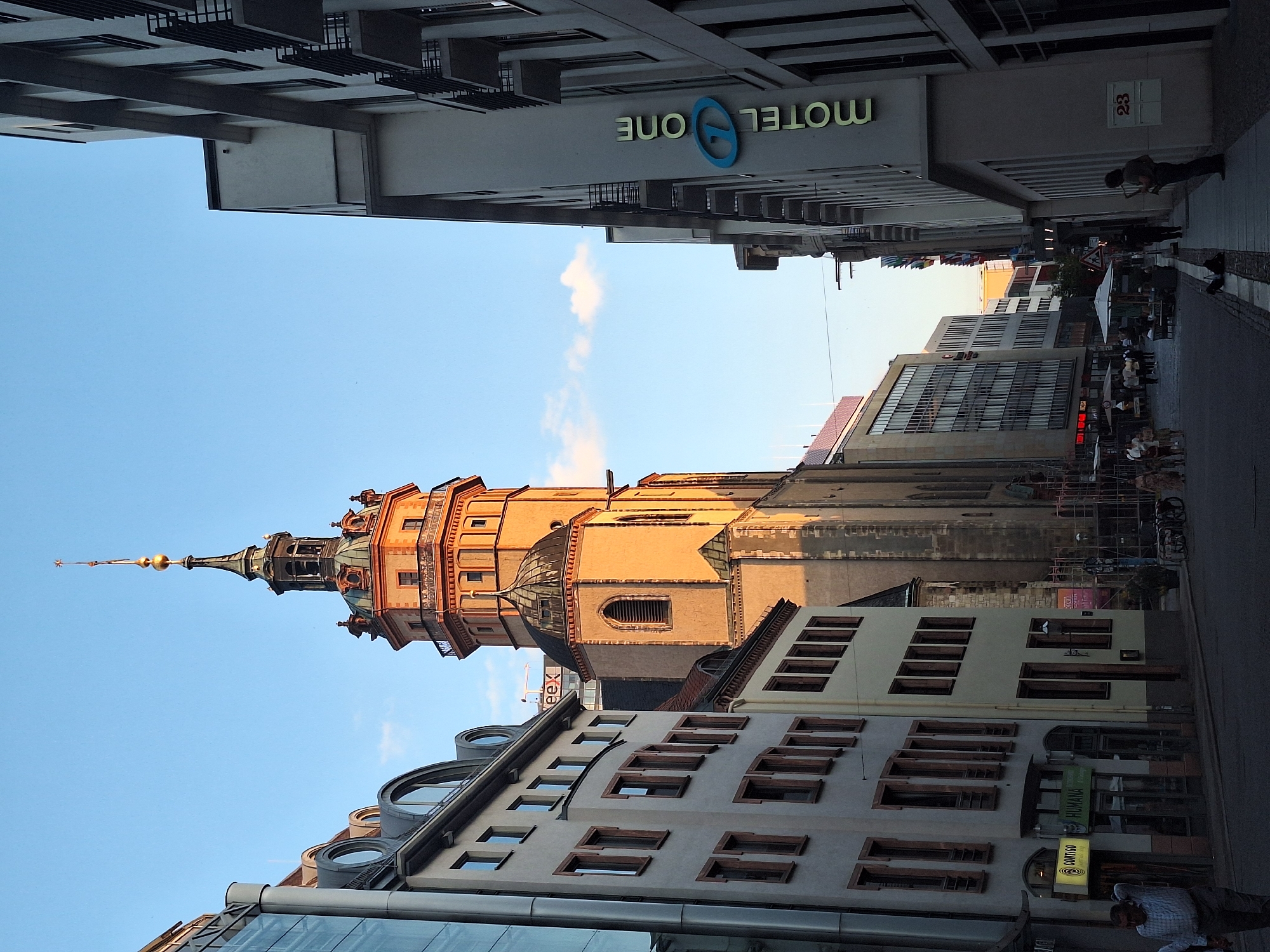 The image captures a street scene in Leipzig, Germany, with a prominent church tower dominating the skyline. The tower, a historical landmark, is bathed in the warm light of the setting sun, casting long shadows on the cobblestone street. Modern buildings with glass facades line the street, creating a contrast between old and new architecture.
Several people are visible in the scene. On the left side of the street, a man in a checkered shirt is walking away from the camera. Further down the street, groups of people are seen dining at outdoor cafes and browsing in shops. On the right side, a lone figure stands on the sidewalk, playing a musical instrument. The street is clean and well-maintained, with parked bicycles and streetlights adding to the urban ambiance. The sky is clear blue, with a few wispy clouds adding texture to the scene. A hotel sign, "MOTEL ONE," is visible on one of the modern buildings. The overall impression is one of a vibrant city that blends its rich history with modern development.