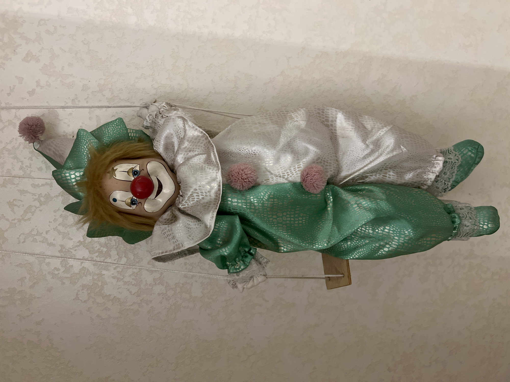 A doll clown is suspended by strings against a textured beige wall. The clown is dressed in a green and white outfit with ruffled collars and cuffs. It has a painted face with blue eyes, red cheeks, and a red nose. A green conical hat with a pink pom-pom sits on its head. The clown is positioned on a small wooden swing.