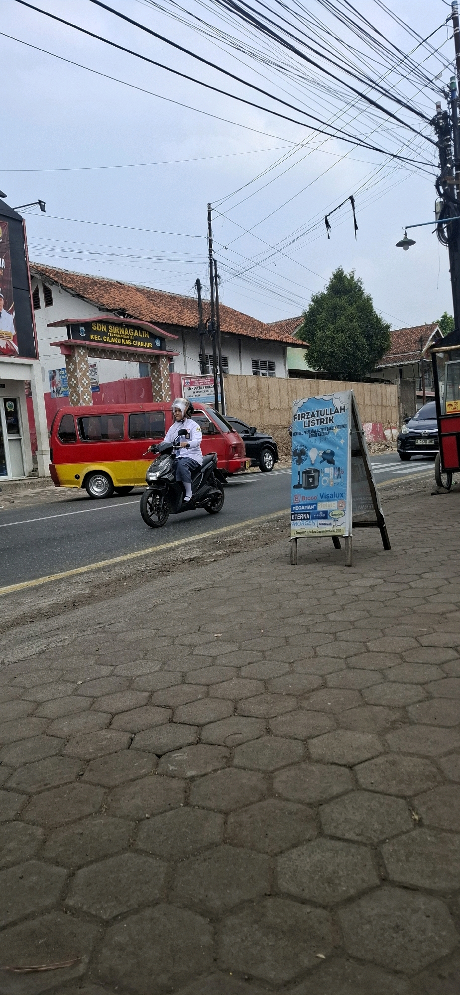 The image captures a street scene in Sirnagalih, Indonesia, featuring a person riding a black scooter on the road. The rider, wearing a white long-sleeved shirt and dark pants, has a mask on. The scooter has a partially visible license plate. To the left of the road, a red and yellow mini-van is parked, and behind it is a building with the inscription "SDN SIRNAGALIH TERAKREDITASI KEC. CILAKU KAB. CIANJUR." A black car is visible further down the road. On the right side of the image, an A-frame sign with Indonesian text, advertisements for electrical products, and brand names like "Visalux" and "Quantum" is standing on a paved sidewalk. The sidewalk is composed of hexagonal concrete pavers. The background shows a building with a red tiled roof, trees, and a complex network of overhead electrical wires. The sky is overcast.