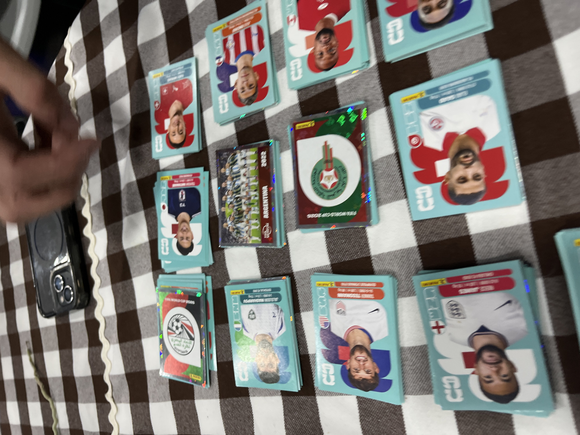 The image shows a collection of Panini World Cup 2022 sticker cards scattered on a tablecloth with a brown and white checkered pattern. Prominent cards include those of national teams such as Argentina, Switzerland, Egypt, USA, and England. One card features the Argentina team photo from the 2022 World Cup. Some cards have individual player portraits. A black smartphone is visible on the left side of the image. A hand partially appears on the left edge of the frame.