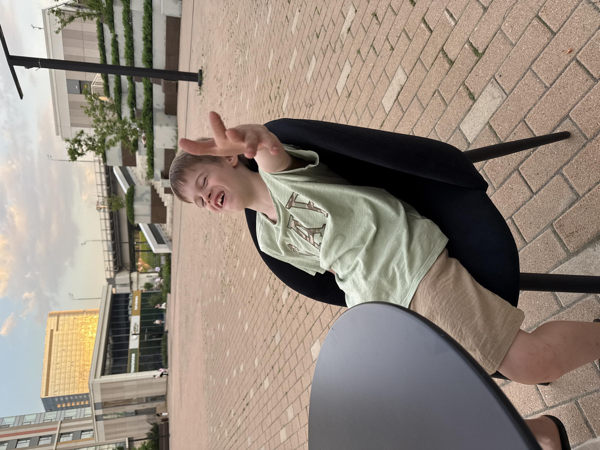 A young boy sits in a black chair at a small, black table, extending his hand towards the camera. The boy wears a light green t-shirt and tan shorts. The scene takes place on a brick-paved area. In the background, there are buildings, including a tall building with a gold-colored facade. Based on the architecture and building styles, this location is likely in Moscow, Russia.