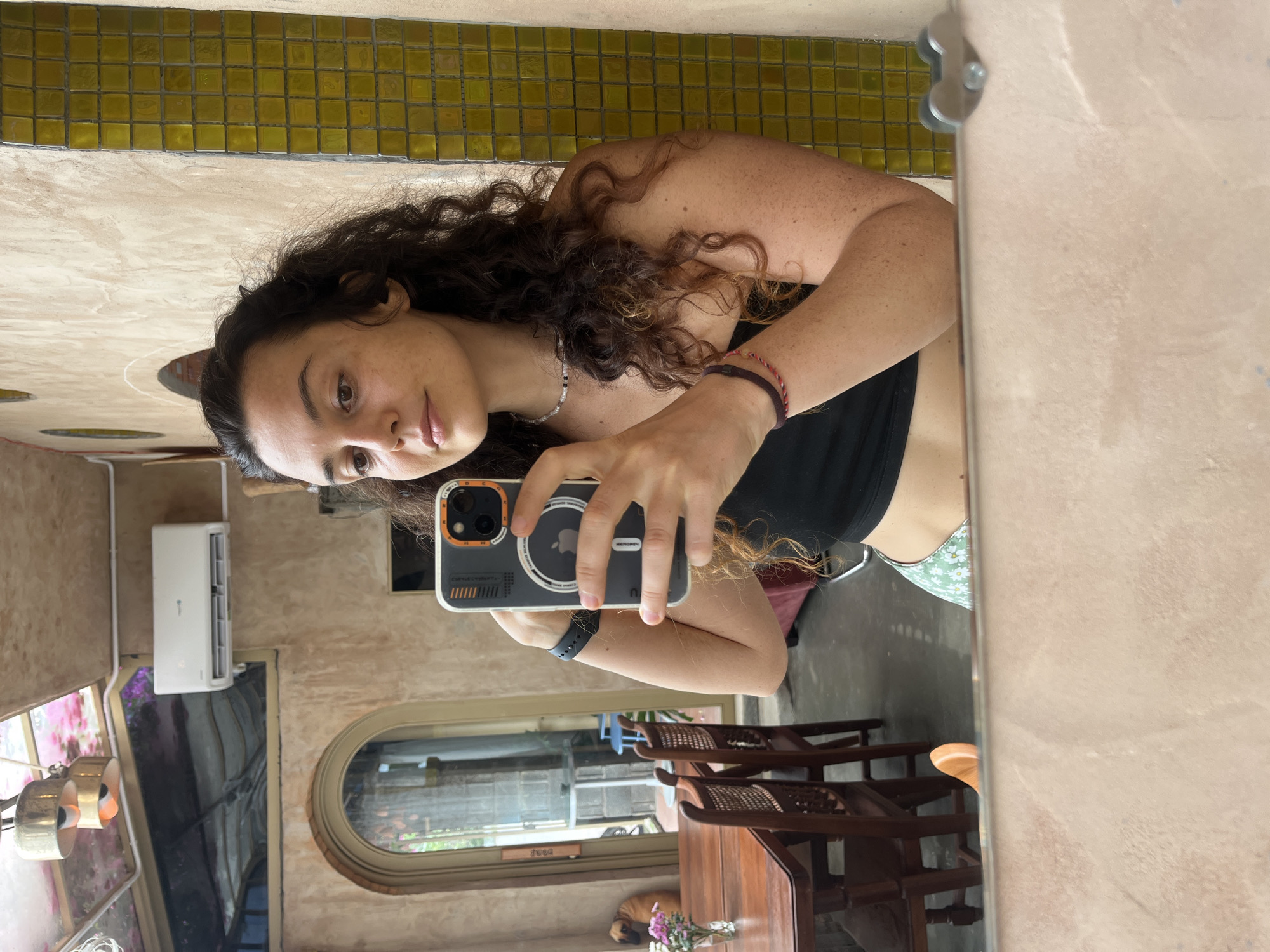 A young woman with long, curly dark hair takes a mirror selfie indoors. She is wearing a black crop top and a light green skirt with a floral pattern. A necklace with small beads adorns her neck, and she wears a dark watch on her left wrist and two bracelets on her right arm. She holds a smartphone in her right hand, angled to capture her reflection. In the background, a dining table with wooden chairs is visible through an arched doorway. A dog sits near the table, and a vase with pink flowers is on the table. The room has textured walls and a tiled accent wall. An air conditioning unit is mounted high on the wall. This scene is located in Mũi Né, Vietnam.