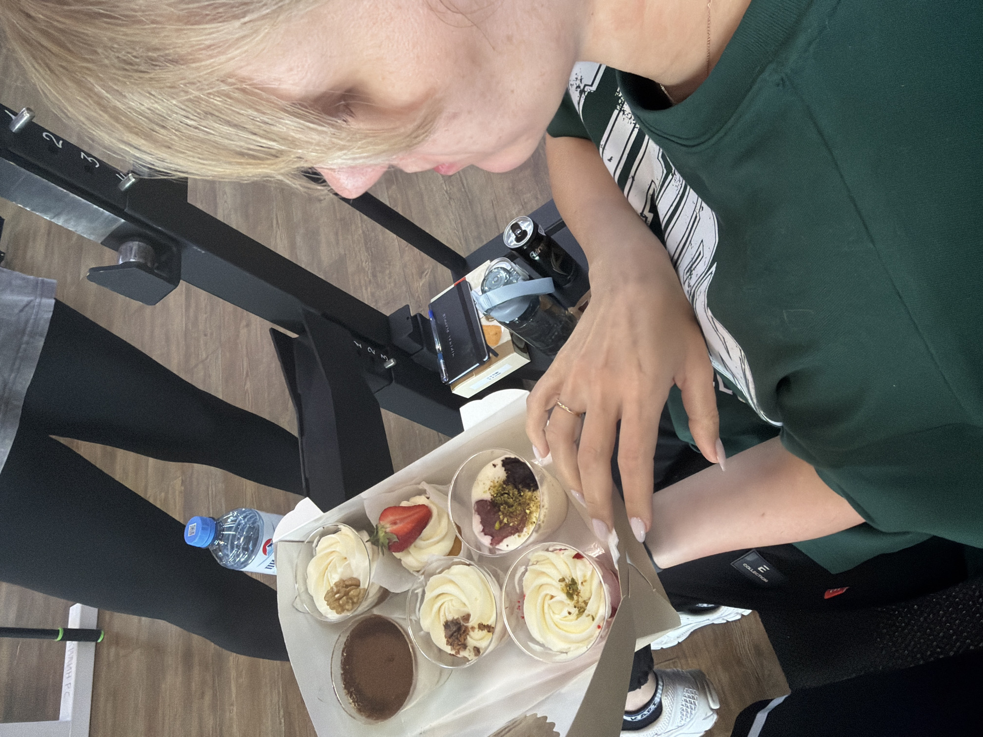 An overhead shot captures a person with blonde hair, seen from the chest up, looking down at a box of desserts. The person is wearing a dark green long-sleeved top with a white graphic on the side, revealing a "PUMA" logo. A thin gold chain is visible around their neck. Their right hand, adorned with a gold ring and light-colored manicured nails, hovers near the desserts in the box.
The white box contains at least six individual desserts in clear plastic cups. These include various parfaits or trifles with cream toppings, some garnished with walnuts, pistachios, strawberry slices, or chocolate powder. One dessert is a solid brown, likely chocolate.
The setting appears to be an indoor space, possibly a gym or fitness studio, indicated by the presence of black exercise equipment with numbered adjustments ("1", "2", "3") in the background. The floor is made of light-colored wood. Another person's legs, wearing black leggings, are partially visible on the left.
Adjacent to the desserts, a blue-capped plastic water bottle with the partially visible label of a Russian brand ("ЭДЕЛЬВЕЙС" / Edelweiss) is on the floor. In the background, on a dark surface, there's a dark notebook with "МОЙ ЖУРНАЛ" (My Journal) written on it, a pen, and a black water bottle with a "PUMA" logo. The person's white sneakers, with a black band featuring "KAPPA" branding, are visible in the lower right. A "COLLECTION" tag is visible on a piece of dark clothing. The scene suggests a break or treat during a fitness-related activity in Chaikovsky, Russia.