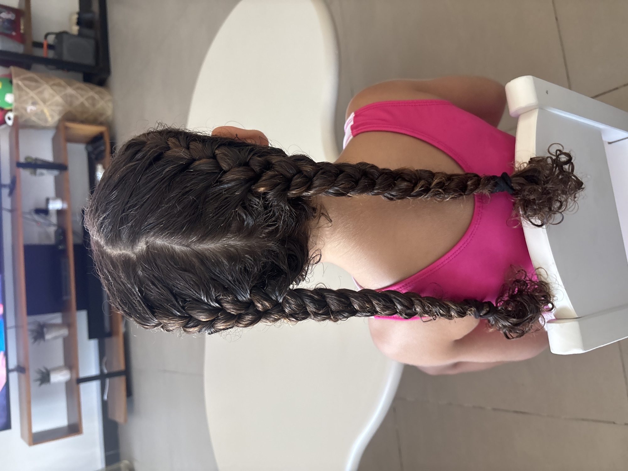 The image shows the back of a young girl with dark brown hair styled in two braids. She is wearing a bright pink sleeveless top and is seated on a white chair, facing away from the camera. The braids are thick and detailed, with the ends slightly curled. The background is blurred, but suggests an indoor setting with a light-colored table or counter in the foreground and a shelf unit with decorative items and electronics in the distance. The floor appears to be tiled. The setting does not contain any discernible indicators of Dubai, United Arab Emirates.