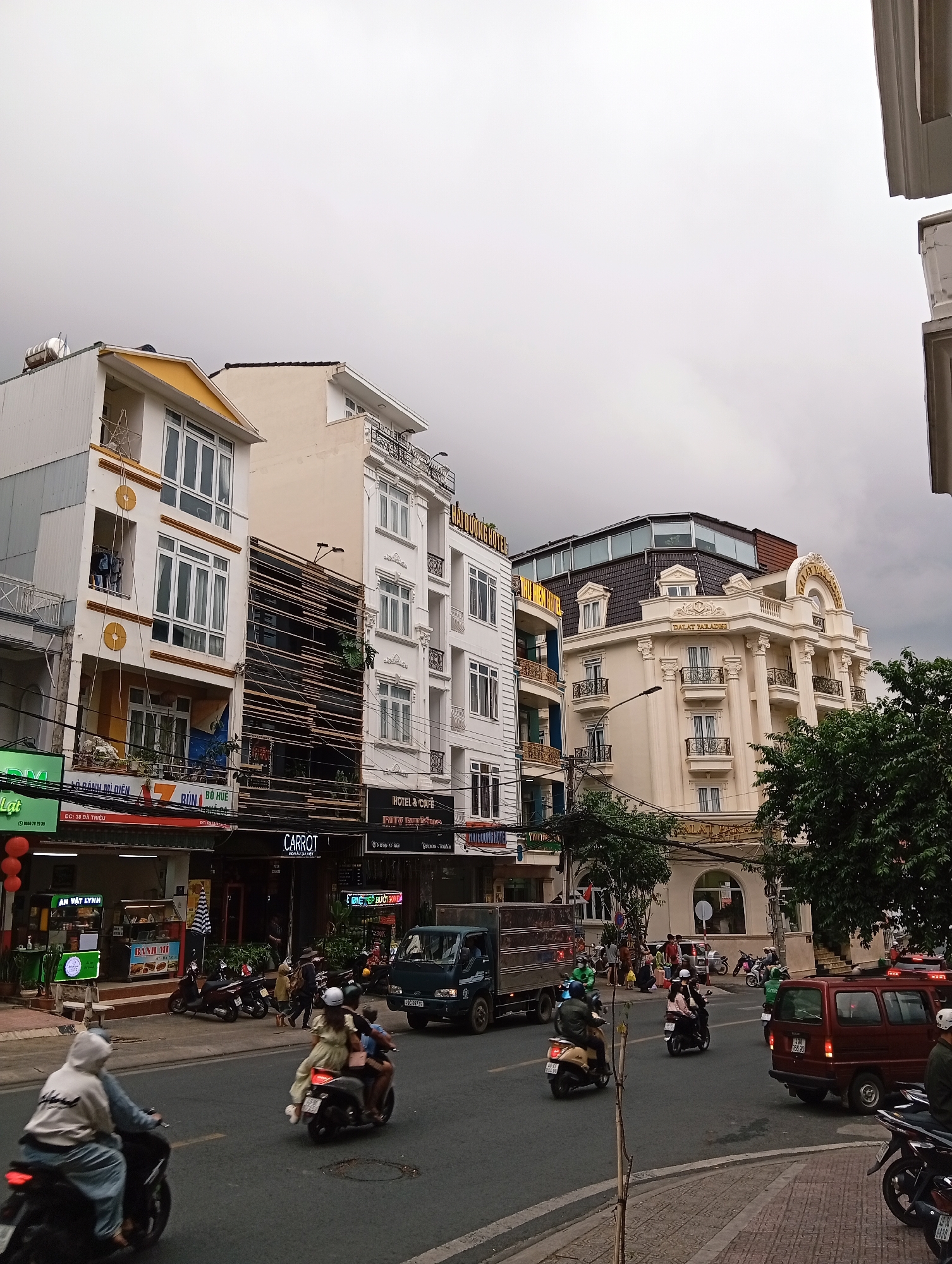 This is a street-level, eye-level, full-shot photograph of a busy street in Da Lat, Vietnam, under a cloudy sky. The street is lined with multi-story buildings, a mix of modern and older architecture. On the left, a tall, narrow building with white and yellow accents features multiple balconies with white railings. Next to it is a darker building with a facade made of horizontal wooden slats, interspersed with some greenery. Further to the right, a cream-colored, ornate building with columns and balconies, identified by signage as "DALAT PARADISE", stands prominently. The ground floor of these buildings houses various businesses, including restaurants and a hotel, with visible signage in Vietnamese.
The street is filled with traffic, primarily motorbikes, which are the dominant mode of transportation. Several individuals are seen riding on motorbikes, some with passengers. A dark blue truck is visible in the middle of the street, and a red minivan is parked on the right side. Pedestrians are also present, walking along the sidewalks and crossing the street. The overall environment suggests a bustling urban commercial area. The presence of multiple businesses, the volume of traffic, and the density of buildings are characteristic of a city center.