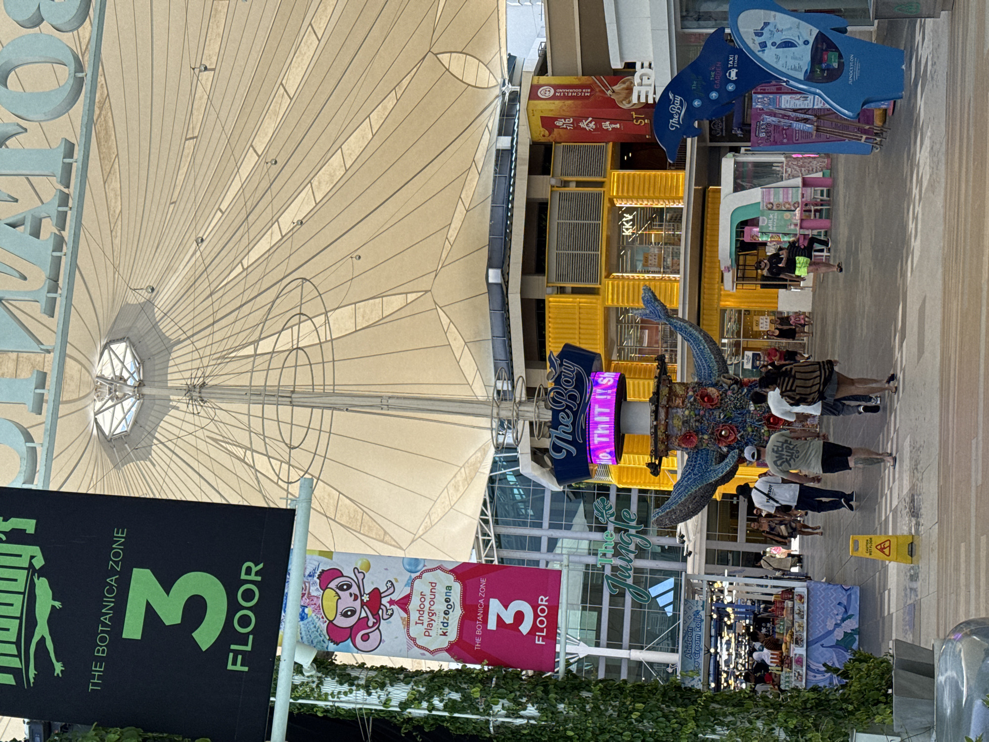 The image depicts an outdoor shopping area in Patong, Thailand, under a large, light-colored tensile fabric canopy. A black banner with green text announces "THE BOTANICA ZONE 3rd FLOOR" and features a cartoon frog logo. Below this, a pink banner shows a cartoon character and advertises "Indoor Playground kidzooona". In the foreground, several people are walking through the open-air mall. To the left, a stall selling ice cream and other items is visible, with shoppers browsing. In the center, a colorful, sculptural totem-like structure stands with a blue whale motif. Beyond this, a bright yellow building with large windows and signage like "KKV" is present. Various shops and stalls line the walkway, and blue signage with shark fins indicates "The Bay" and a "Taxi Stand". Green ivy climbs a tall pillar on the far left. A yellow "Caution Wet Floor" sign is visible on the ground. The overall scene is bright and appears to be during daylight hours.