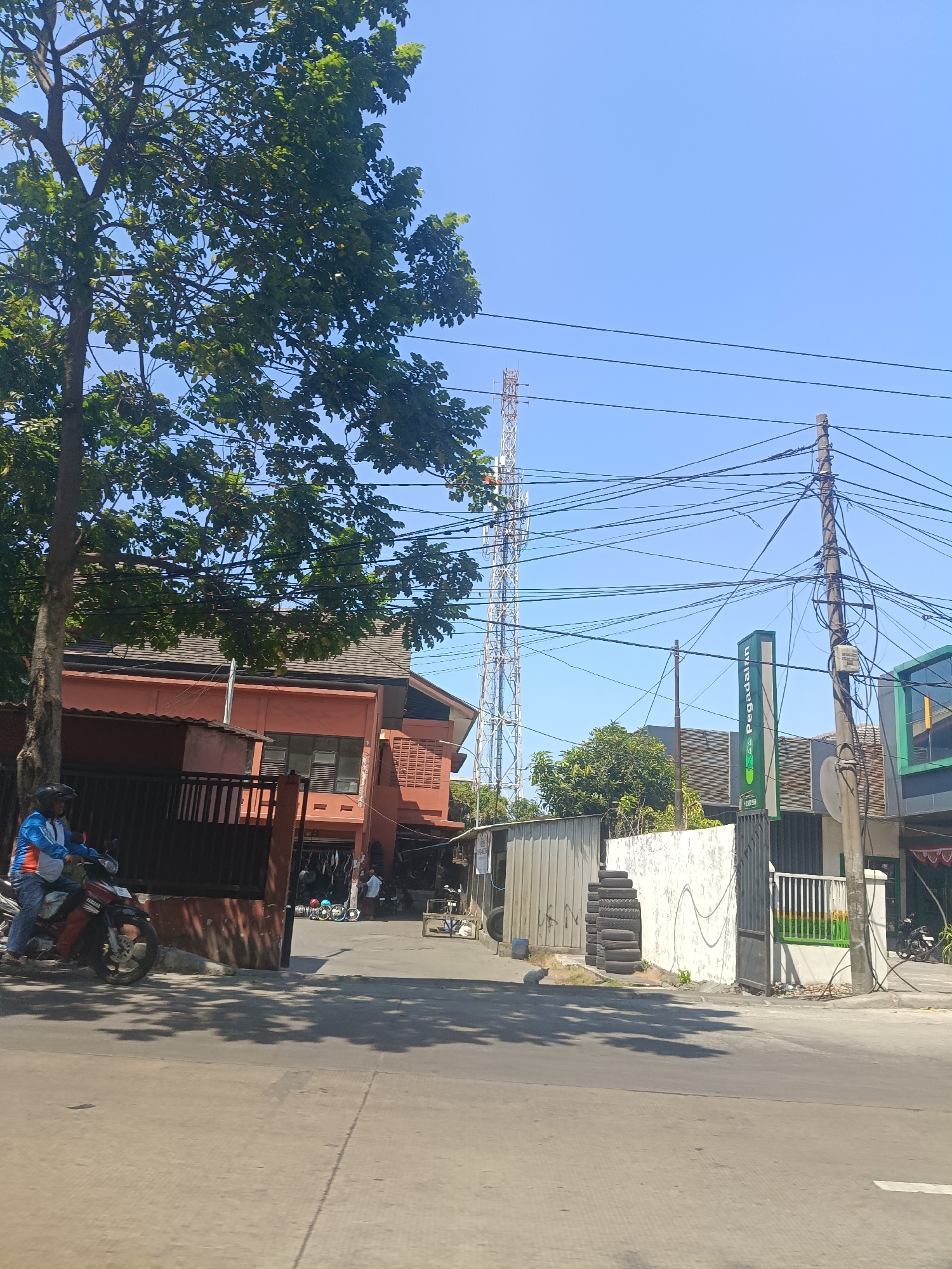 A street scene in Surabaya, Indonesia, bathed in bright sunlight under a clear blue sky. The foreground shows a gray asphalt road with dappled shadows from overhead trees. A motorcycle with a rider wearing a blue and white jacket and jeans is parked on the left side of the road. To the right of the road, a building with an orange facade and dark brown window frames is visible. Behind this building, a tall, lattice telecommunication tower stands prominently against the sky. Further along the street, a white wall with a green sign that reads "Pegadaian" is present. A concrete utility pole with numerous wires is situated in front of this wall. A stack of car tires is piled next to the white wall. Various other buildings and a smaller telecommunication pole are also visible in the background, along with more power lines crisscrossing the scene. The overall impression is of a typical urban street environment in a commercial or mixed-use area.