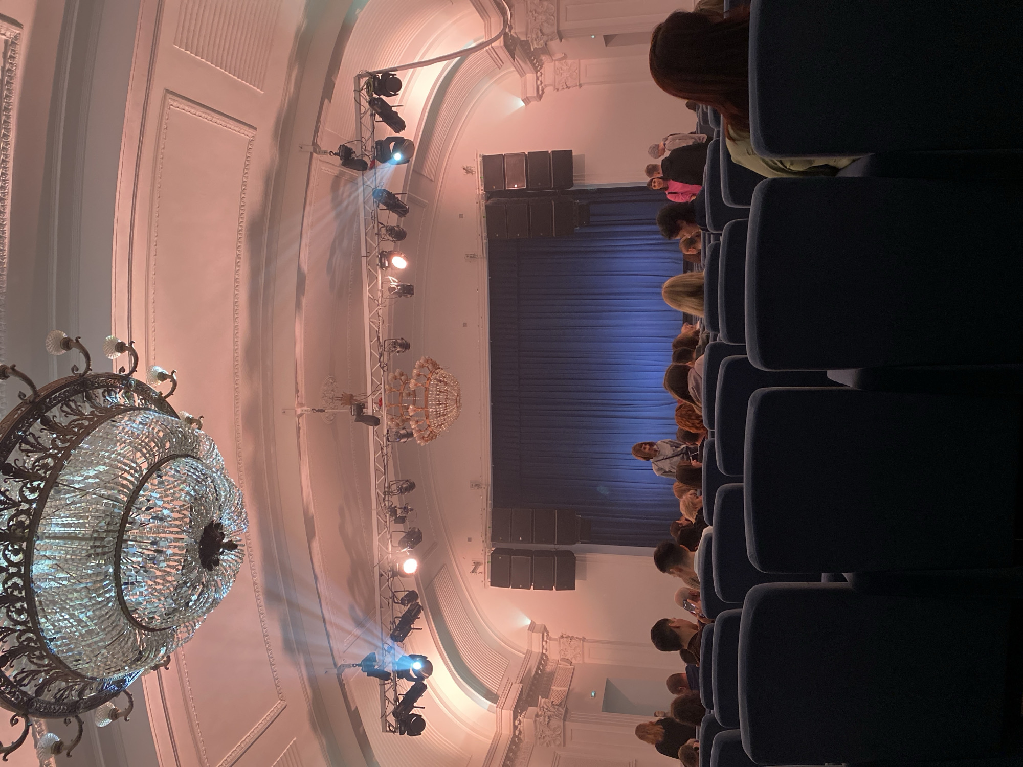 The image shows the interior of a theater. Rows of dark blue chairs are visible in the foreground, occupied by an audience of people. The stage features a blue curtain backdrop and a stack of speaker boxes above it. A metal truss holding stage lighting is visible on each side of the stage. Two large chandeliers hang from the ceiling, which is decorated with ornate moldings. The image captures the ambiance of a theatrical performance, likely in a historical or grand venue. Based on the architecture, this could potentially be a theater or concert hall in the city of Kazan, Russia, though confirmation would require additional visual or contextual clues.