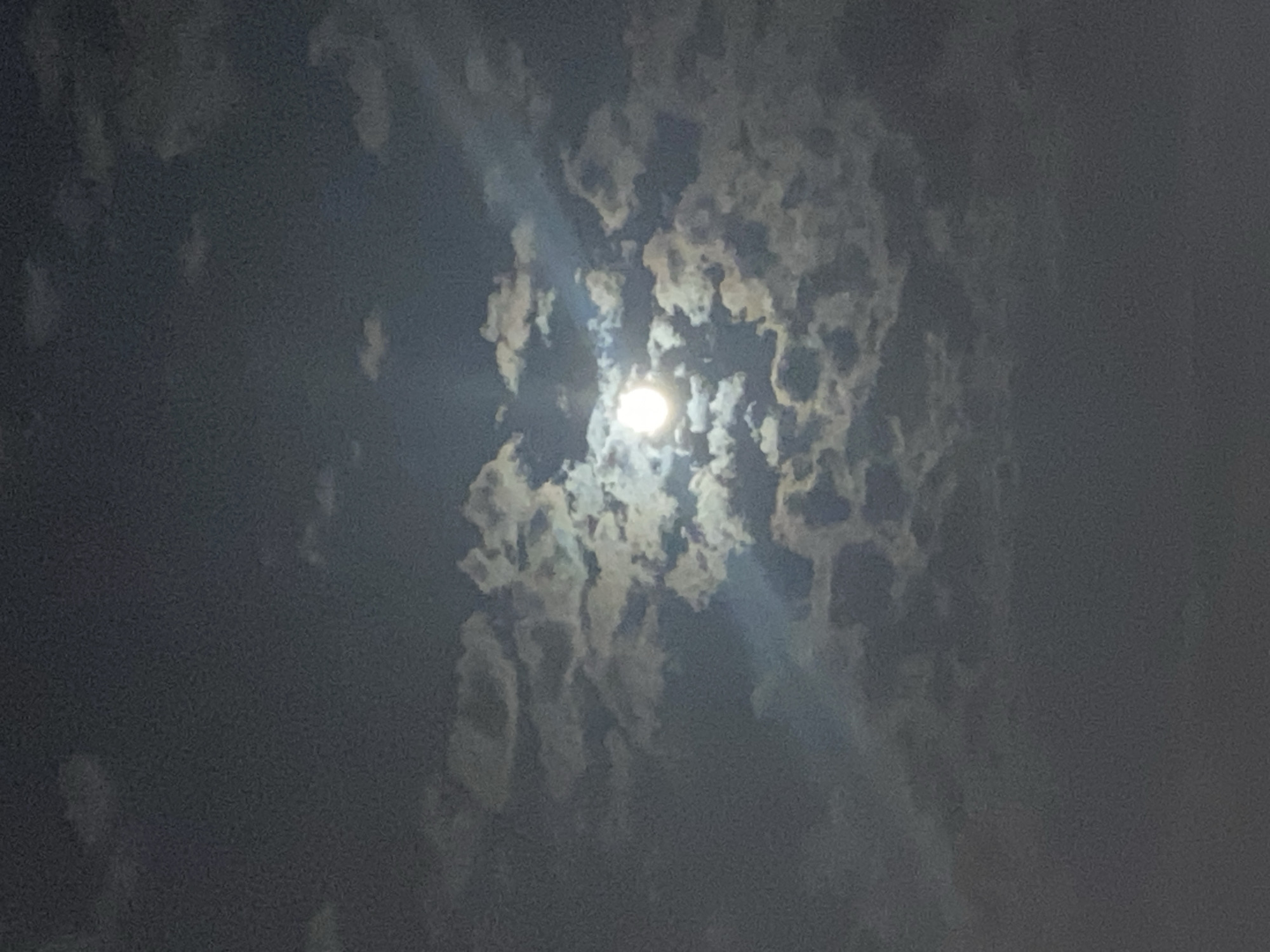 The image shows a full moon partially obscured by clouds at night. The moon is bright and appears as a white disc, with some of its light illuminating the edges of the surrounding clouds, creating a halo effect. The clouds are varied in density, with some appearing wispy and thin, while others are thicker and more textured, casting shadows that add depth to the scene. A faint beam of light emanates from the moon, cutting through the clouds at an angle. The overall sky is a deep, dark blue-gray, indicative of nighttime. There are no visible landmarks or structures to suggest a specific location in Iași, Romania, or any city. The image is solely focused on the celestial display.