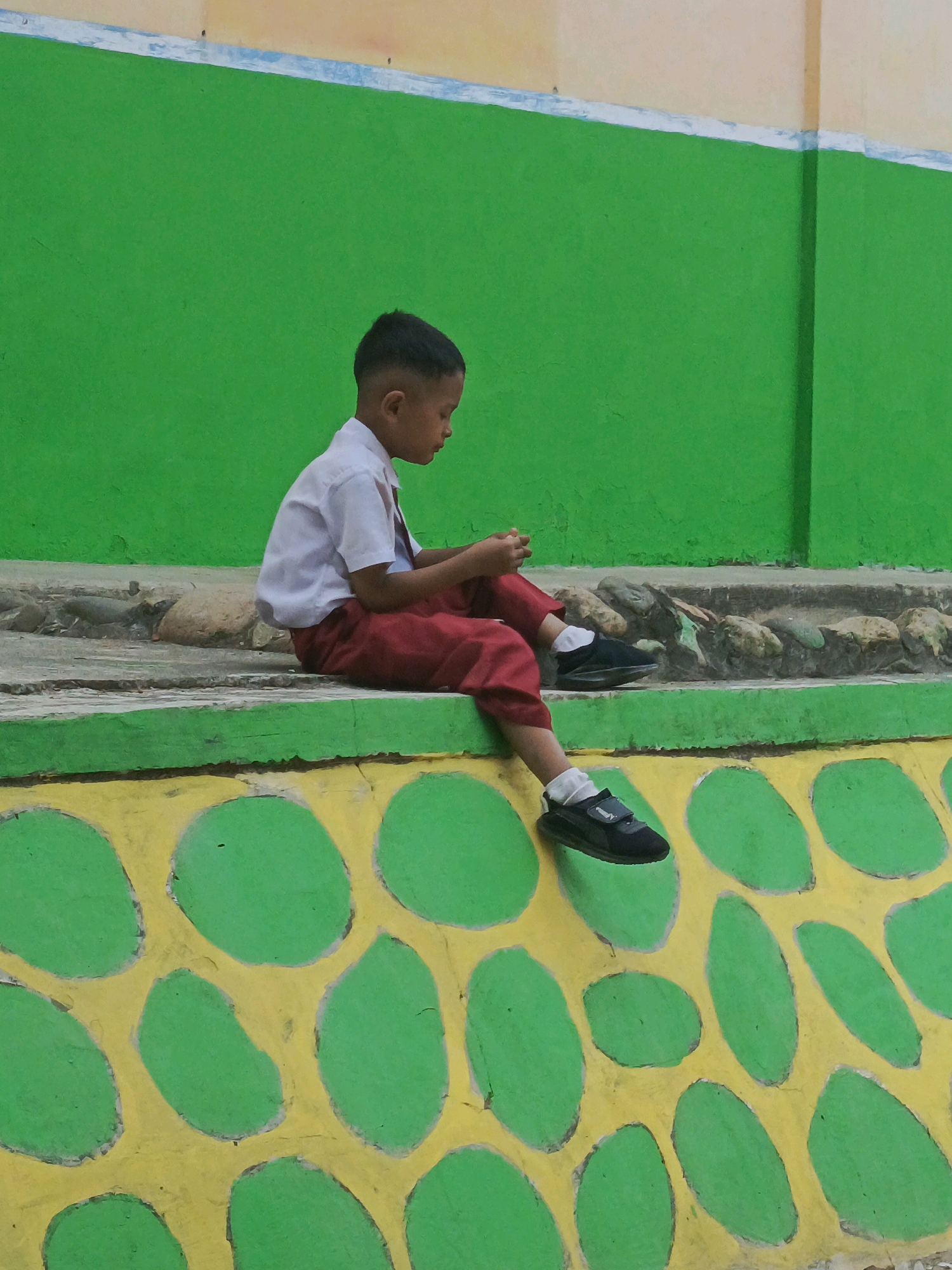 A young boy in a white shirt and red pants sits on a concrete ledge. The boy is facing to the right and looking down at his hands. He is wearing black shoes and white socks. Behind him is a bright green wall, and in front of him is a yellow and green patterned wall. The scene appears to be outdoors in an urban setting. The location is Karya Dadi, Indonesia.