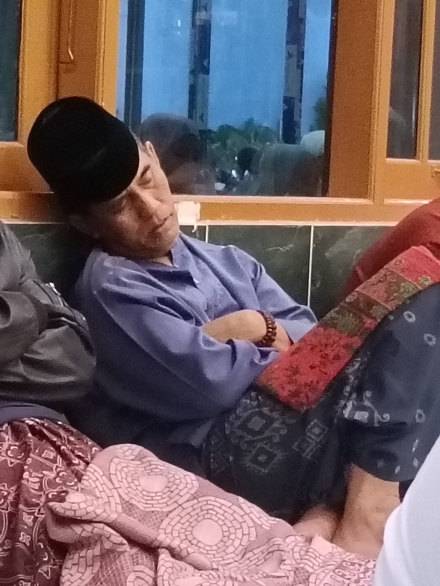 A medium shot shows a man wearing a black peci hat, a light blue long-sleeved shirt, and patterned pants. He is leaning his head against a wall or window frame, with his eyes closed and his arms crossed over his chest. A beaded bracelet is visible on his right wrist. The man appears to be resting or sleeping. To his left, another person's shoulder and arm are visible, draped in dark clothing. The background shows a window with a view of blurred greenery outside, framed by a wooden structure. The setting suggests an indoor environment, possibly a home or a place of worship, in Kediri City, Indonesia. The lighting is dim, indicating it could be late evening or early morning. The overall mood is peaceful and quiet.
