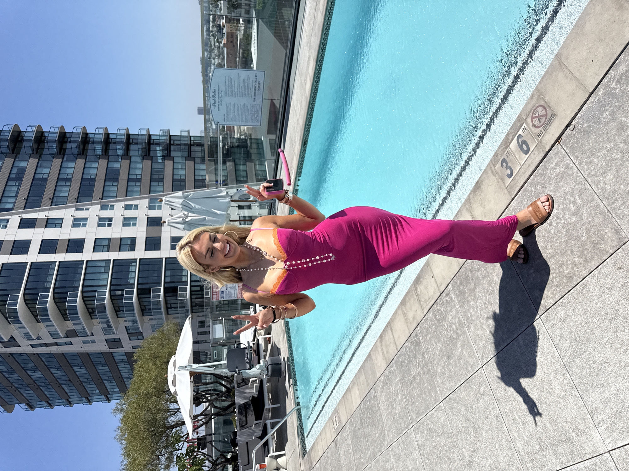 The image shows a woman in a pink dress standing by a pool on a rooftop with a cityscape in the background. The pool has a no diving sign and depth markings. The woman is posing with a peace sign, holding a phone, and wearing jewelry. The location is a rooftop pool in Los Angeles, California, as indicated by the presence of the Godfrey Hotel in the background. Pool rules are printed on a board in the background. The woman is wearing sandals.