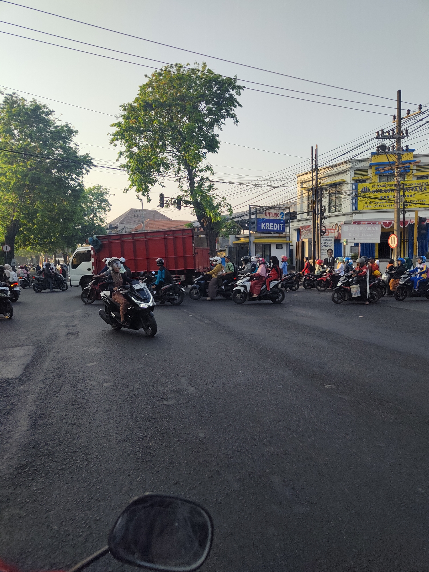 The image depicts a busy street intersection in Bangil, Indonesia, likely during daylight hours. The foreground shows a portion of a motorcycle's rearview mirror and handlebars, suggesting the photo was taken from a moving motorcycle. The road surface is asphalt, with some visible cracks and wear.
Numerous motorcycles are present, with many riders wearing helmets. People are seen on the motorcycles, some of them appear to be traveling in a convoy or group. One white truck with a red cargo bed is stopped at the intersection, with a few people standing on its rear.
To the right of the intersection, a yellow and white building with signage is visible. One sign has the words "PT. BANK PERKREDITAN RAKYAT" and "SURAKARTA UTAMA KANTOR PUSAT" printed on it. Another sign displays "KLINIK AMANI". Traffic lights are also present at the intersection. Trees line the sides of the road, casting shadows on the street. The sky is a pale blue, indicating clear weather. The overall scene suggests moderate traffic and daily life in an urban Indonesian setting.