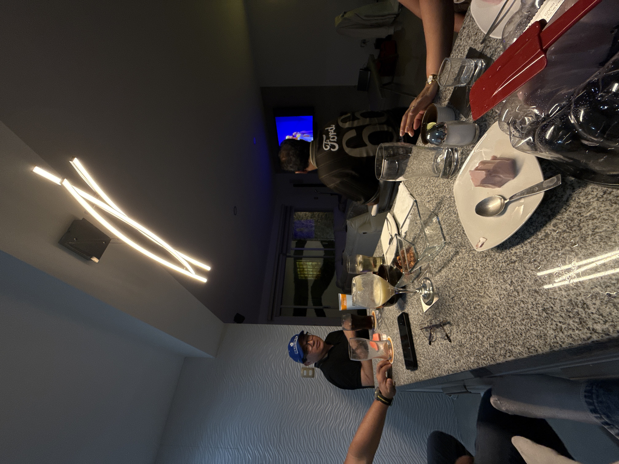 Here's a description of the image:
**Scene:** An indoor gathering, possibly a dinner or party, in a modernly decorated room.
**Location:** Santa Clara Ocoyucan, Mexico
**People:**
* Several individuals are present, including at least one man wearing a blue baseball cap and glasses, one man wearing a jersey with "Ford 55" on the back, and at least two other people who are partially visible.
* One person has a watch on their wrist.
**Objects:**
* A countertop, likely in a kitchen or bar area, is covered with food, plates, glassware, and serving utensils.
* Dishes include what appears to be a plate with meat.
* Various types of glasses are present, some filled with beverages.
* A red knife is visible.
* A television is visible in the background, displaying a blue screen.
* A remote control is lying on the countertop.
* A modern lighting fixture is mounted on the ceiling.
**Actions & Interactions:**
* People are gathered around the countertop, suggesting they are interacting or preparing to eat.
* Someone is pouring a drink.
**Environment:**
* The room has modern decor, with a textured wall and contemporary lighting.
* It appears to be evening or nighttime.
* A window with partially drawn blinds can be seen in the background.