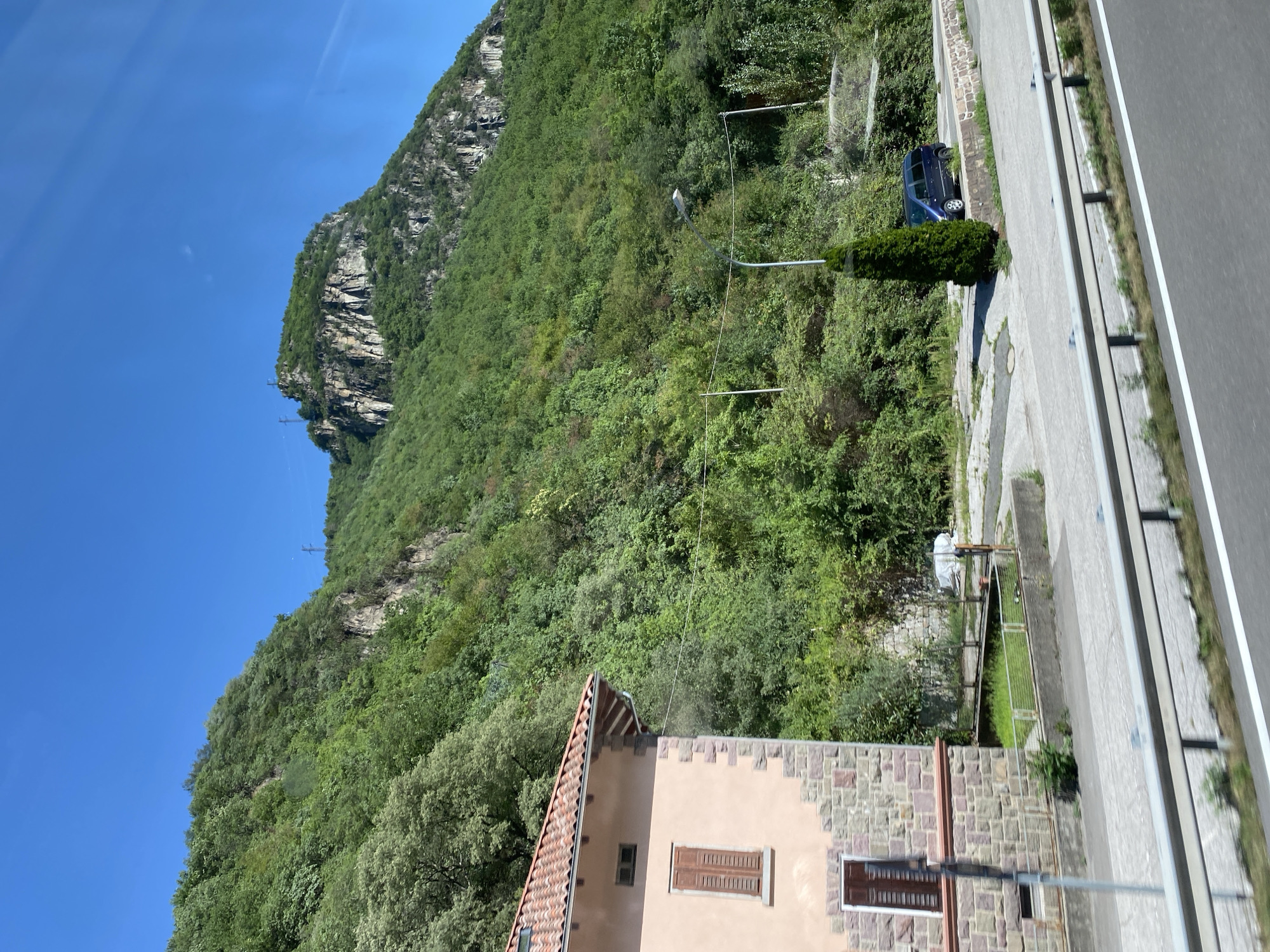 The image shows a view from a moving vehicle on a sunny day, looking towards a mountainous landscape. In the foreground, a light pink building with a tiled roof and stone accents is visible on the left. The building has at least two windows, one with brown shutters. A silver guardrail runs horizontally across the lower part of the frame, indicating the edge of a road. A dark blue minivan is parked on the right side of the road, next to a tall, conical evergreen tree. Behind the minivan, there is a steep, tree-covered mountainside. The mountain features rocky outcrops at its peak, and power lines are visible against the clear blue sky. The environment appears to be rural or semi-rural, with lush vegetation covering the slopes. The scene is likely situated in the Ritten-Renon area of Italy, given the architectural style of the building and the mountainous terrain.