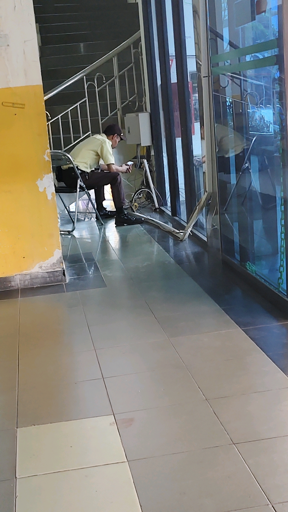A person is sitting on a folding chair in what appears to be a public indoor space. The person is wearing a light-colored short-sleeved shirt, dark pants, and a dark baseball cap. They are holding a mobile phone in their hands and looking down at it, suggesting they are engaged with the device. Beside them, to the right, is a bicycle with its kickstand down, leaning against a wall. The floor is tiled in a neutral color. Behind the person, a staircase with a metal railing ascends. To the right of the staircase, a white electrical box is mounted on the wall. The overall environment suggests a transit area or a communal space within a building. The specific location in Banjarmasin, Indonesia, is not explicitly determinable from the visual cues in the image.