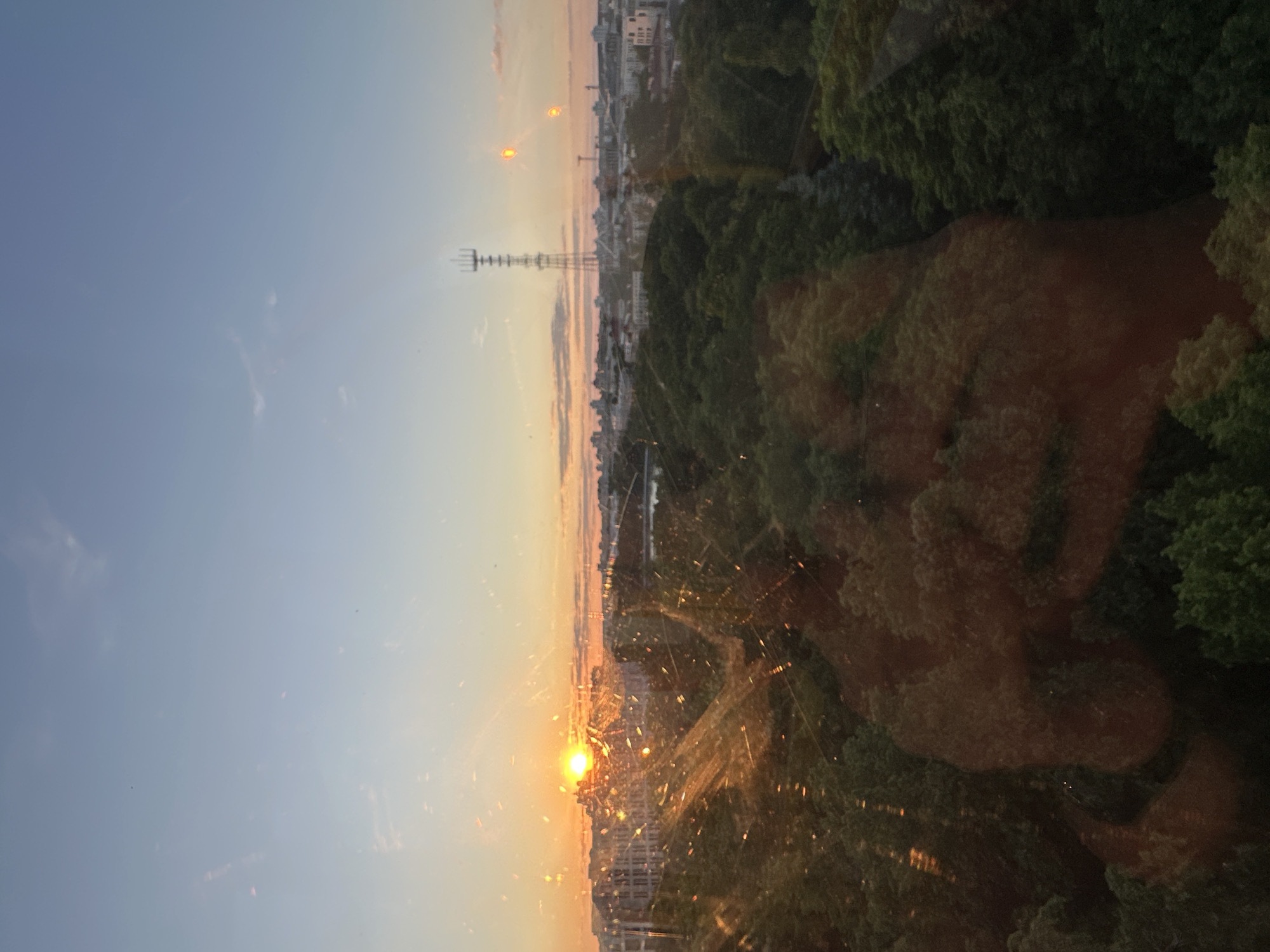 A panoramic view captures a sunset over the city of Minsk, Belarus, as seen from an elevated perspective, likely through a window. The sky dominates the upper left, transitioning from a clear, pale blue to warm orange and yellow hues near the horizon where the sun is setting. The sun itself is a bright, intense orb of light, positioned low on the left, casting a radiant glow.
Along the horizon, the silhouettes of numerous city buildings stretch across the mid-ground. A tall, prominent lattice-structure telecommunications or TV tower, identifiable as the Minsk TV Tower, stands vertically in the center-left, piercing the sky. Below the cityscape, extensive areas of dense green trees cover the right and lower parts of the frame, suggesting the presence of parks or forested areas within the city.
The image appears to have been taken through a window, as evidenced by visible specks of dust and dirt, particularly illuminated by the sunlight, creating starburst effects around the sun. Additionally, a faint, semi-transparent reflection resembling a human face or upper body is visible within the dark green foliage on the right side of the image. The time of day is clearly sunset, indicated by the low sun and the warm, soft light bathing the scene. There is no visible text in the image.
