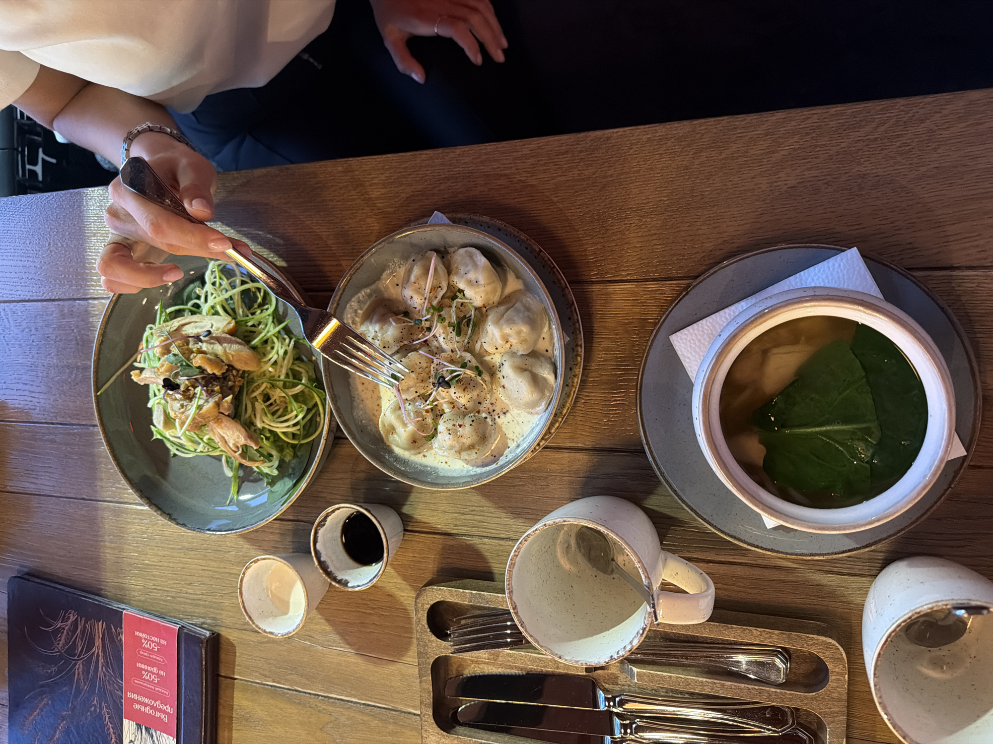 The image captures a top-down view of a meal being enjoyed at a wooden table in what appears to be a restaurant or cafe in Krasnoyarsk, Russia. The setting is indoors, with warm, dim lighting suggesting an evening meal.
In the upper left, a person, visible from the chest down, wears a light-colored top and dark trousers. Their left hand, adorned with a thin ring and a bracelet, holds a fork, poised to pick food from a bowl. The nails are neatly manicured.
Three main dishes are presented on the table. To the left, a grey-green bowl contains a vibrant green salad, possibly made with zucchini noodles or similar vegetable pasta, topped with pieces of what looks like fried or roasted chicken or duck and garnished with microgreens and sesame seeds. In the center, another grey-green bowl holds several dumplings, possibly pelmeni or vareniki, swimming in a creamy white sauce and sprinkled with pepper and microgreens. To the right, a ceramic bowl sits on a grey plate, containing a clear broth or soup with a large green leaf, likely spinach or sorrel, floating on its surface.
The table is set with various other items. Two small, rustic-looking ceramic cups are present, one holding a dark liquid like soy sauce, the other empty. Two larger, speckled ceramic mugs are also visible, one standing upright, the other tipped over, next to a wooden tray holding an assortment of silver cutlery, including knives, forks, and spoons. A dark-covered menu book lies on the table's left side. A red strip on the menu prominently displays Russian text: "ВЫГОДНЫЕ предложения" (ADVANTAGEOUS offers), followed by "-50% НА НАСТОЙКИ" (50% OFF TINCTURES/INFUSIONS) and "-50% НА ДРАНИКИ" (50% OFF DRANIKI - potato pancakes).