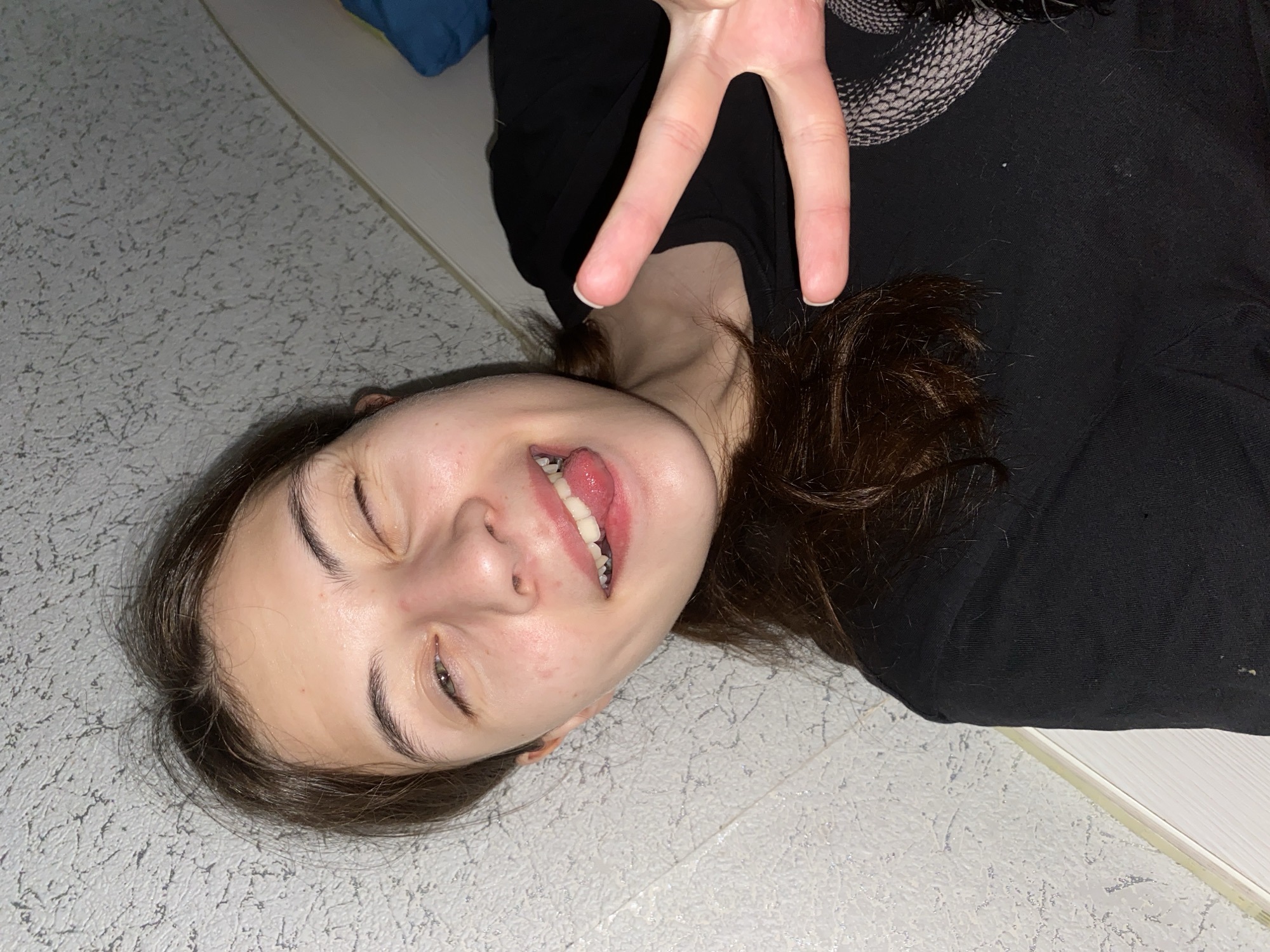 A young female is taking a selfie in a room. She has long, dark brown hair pulled back in a ponytail. Her eyes are closed in a wink, and her tongue is sticking out with her mouth open in a smile. She is wearing a black t-shirt. Her right hand is held up to the camera, with her index and middle fingers extended in a peace sign. The background appears to be a textured, light-colored wall. There is no discernible location in Votkinsk, Russia.