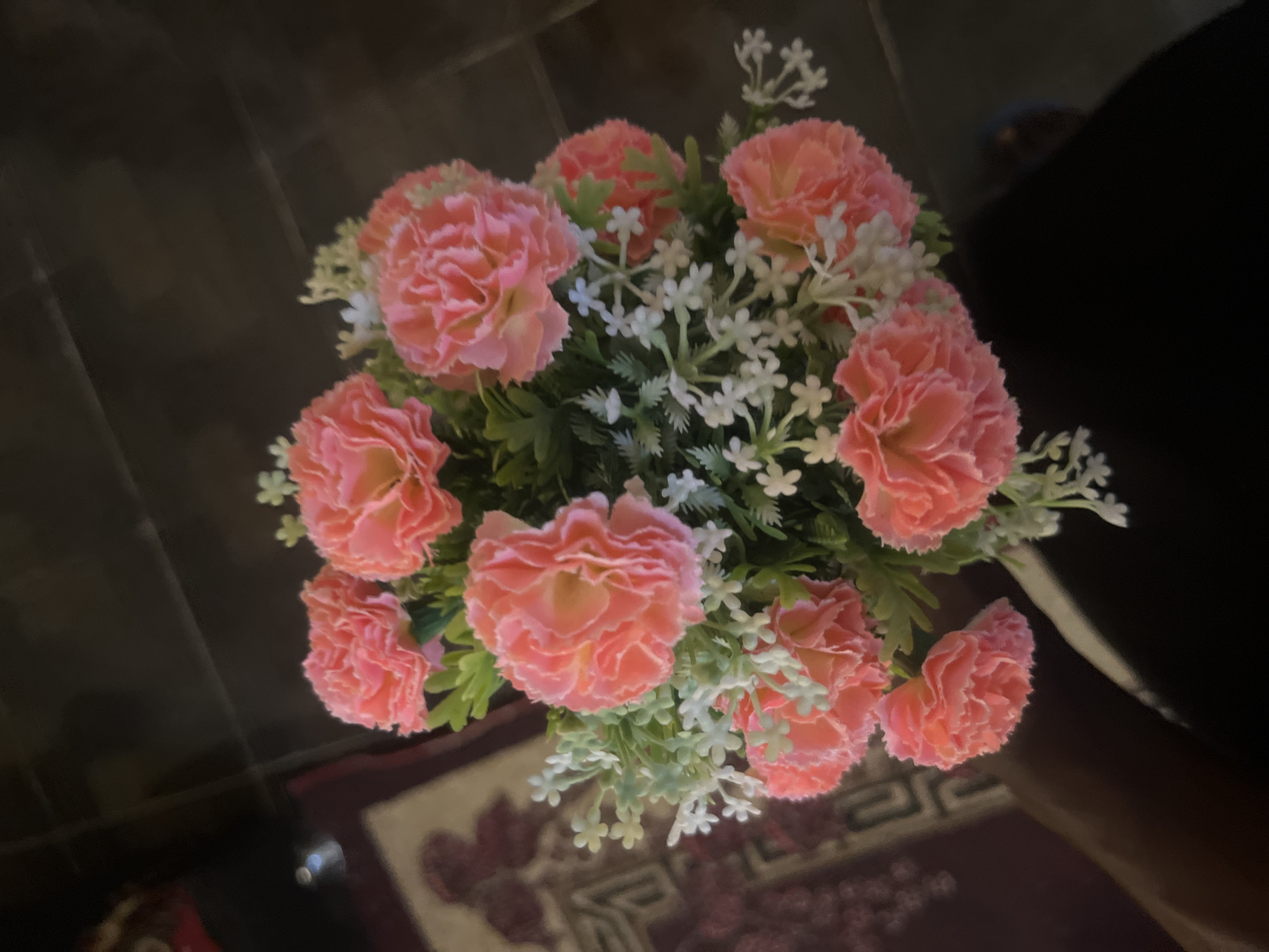 The image shows a close-up, overhead view of a bouquet of artificial pink carnations with small white filler flowers and green leaves. The bouquet is arranged in a round shape and appears to be placed on a dark surface, possibly a table or floor. In the background, a portion of a patterned rug or carpet is visible, with dark, out-of-focus elements suggesting a dimly lit indoor environment. There are no people or specific landmarks visible that would indicate a location in the city of Barakin Akawo, Nigeria.